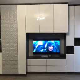 KOCHACZEWSKA DESIGN ANNA KOCHACZEWSKA-LIS - Biała zabudowa meblowa z błyszczącymi frontami, telewizorem w centralnej części i dekoracyjną ścianką z geometrycznym wzorem 3D po lewej stronie.