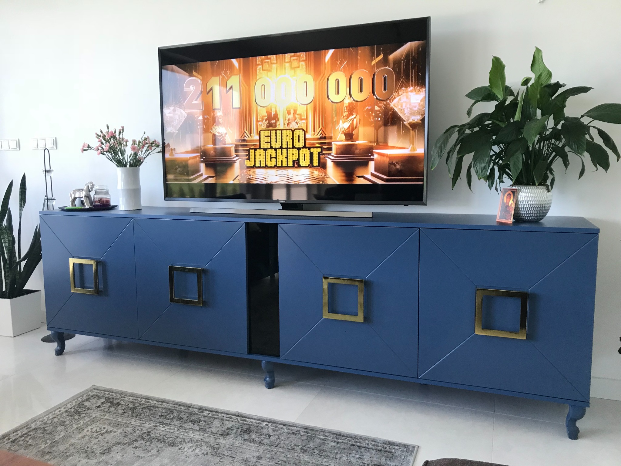 Niebieska szafka RTV z czterema drzwiami z kwadratowymi, złotymi uchwytami, stojąca na małych nóżkach, na której stoi telewizor z włączoną grą Euro Jackpot oraz dekoracje w postaci kwiatów i roślin...