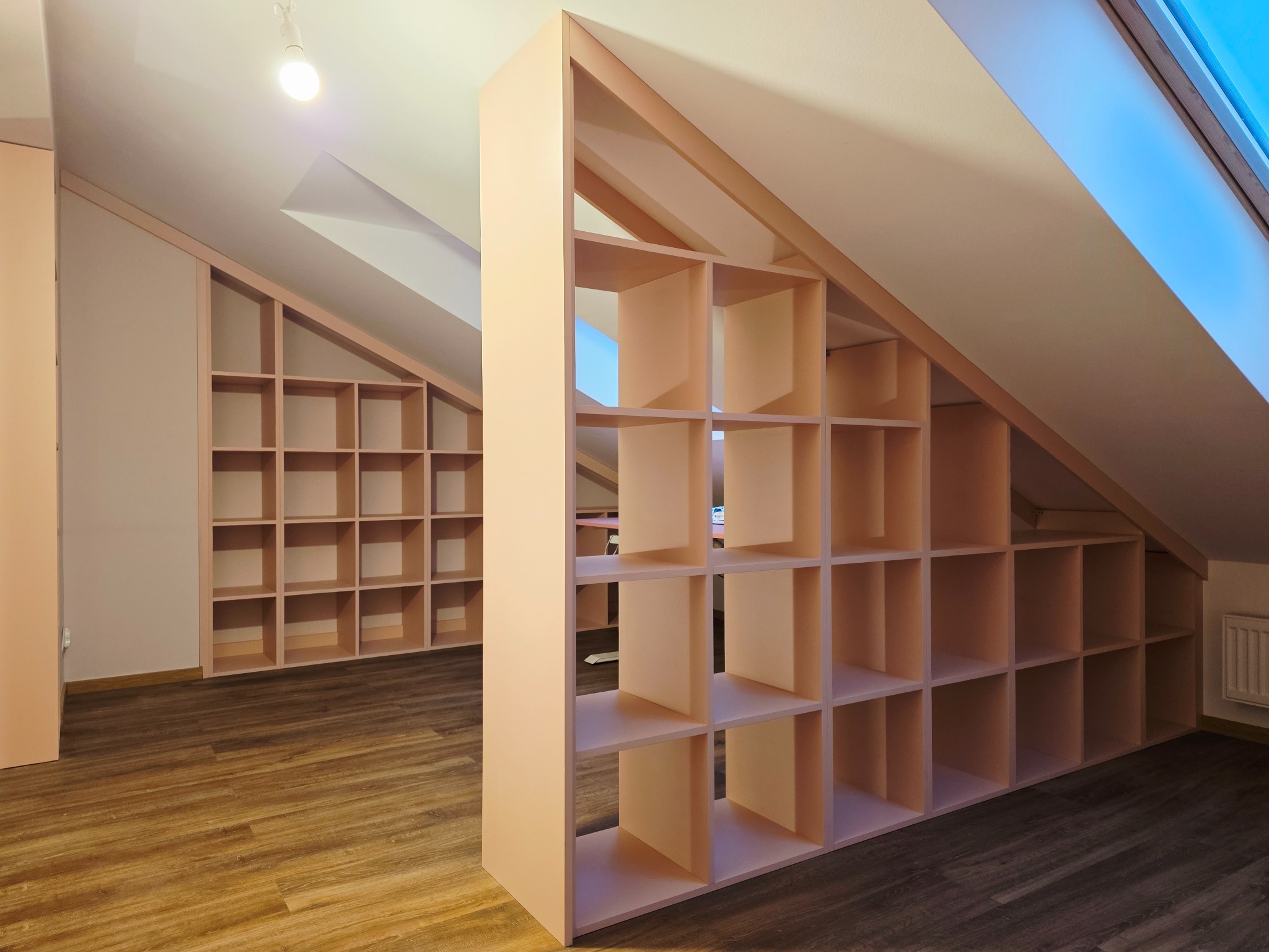 Regał na poddaszu w odcieniach beżu, z licznymi półkami tworzącymi kwadratowe przegródki, dopasowany do skosu dachu. Widoczne drewniane panele podłogowe i okno dachowe.
