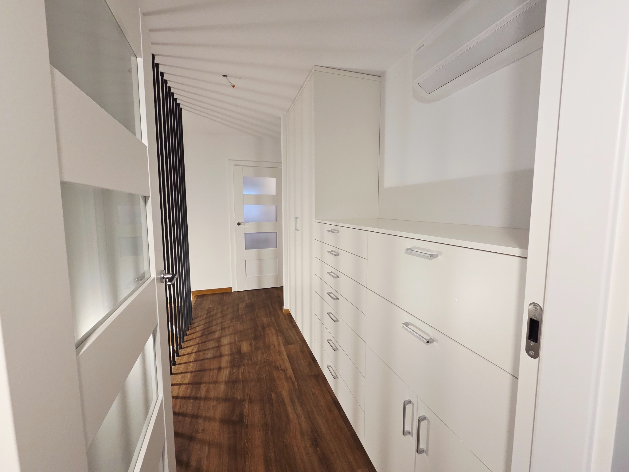 Biała szafa wnękowa z szufladami i szafkami, widok z perspektywy korytarza. Realizacja w Warszawie, minimalistyczny design, drewniana podłoga.