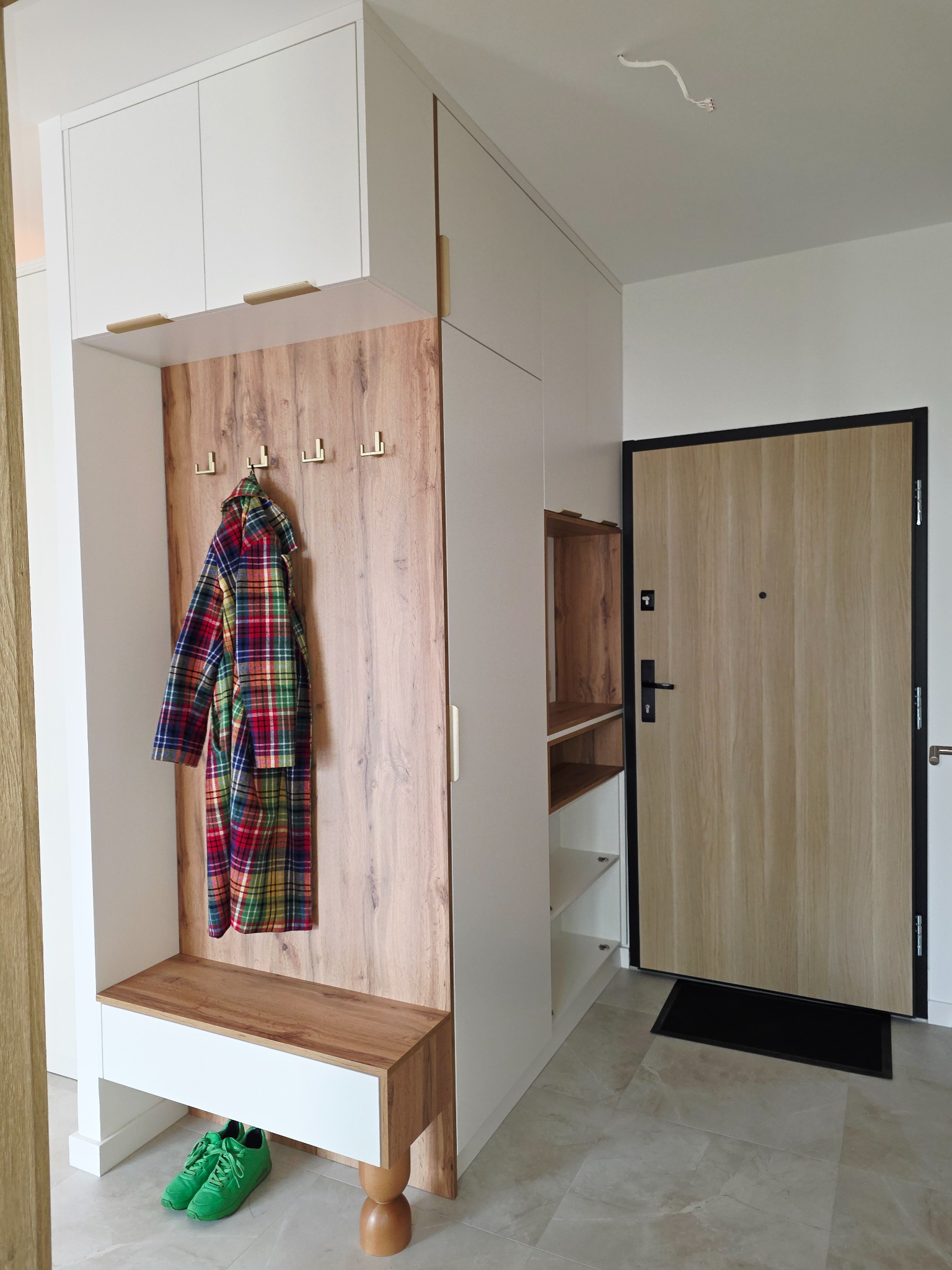 Funkcjonalna zabudowa przedpokoju z szafkami, wieszakami i siedziskiem z drewnianym panelem. Kolorowa kurtka i zielone buty dodają akcentu. Drzwi wejściowe w tle.