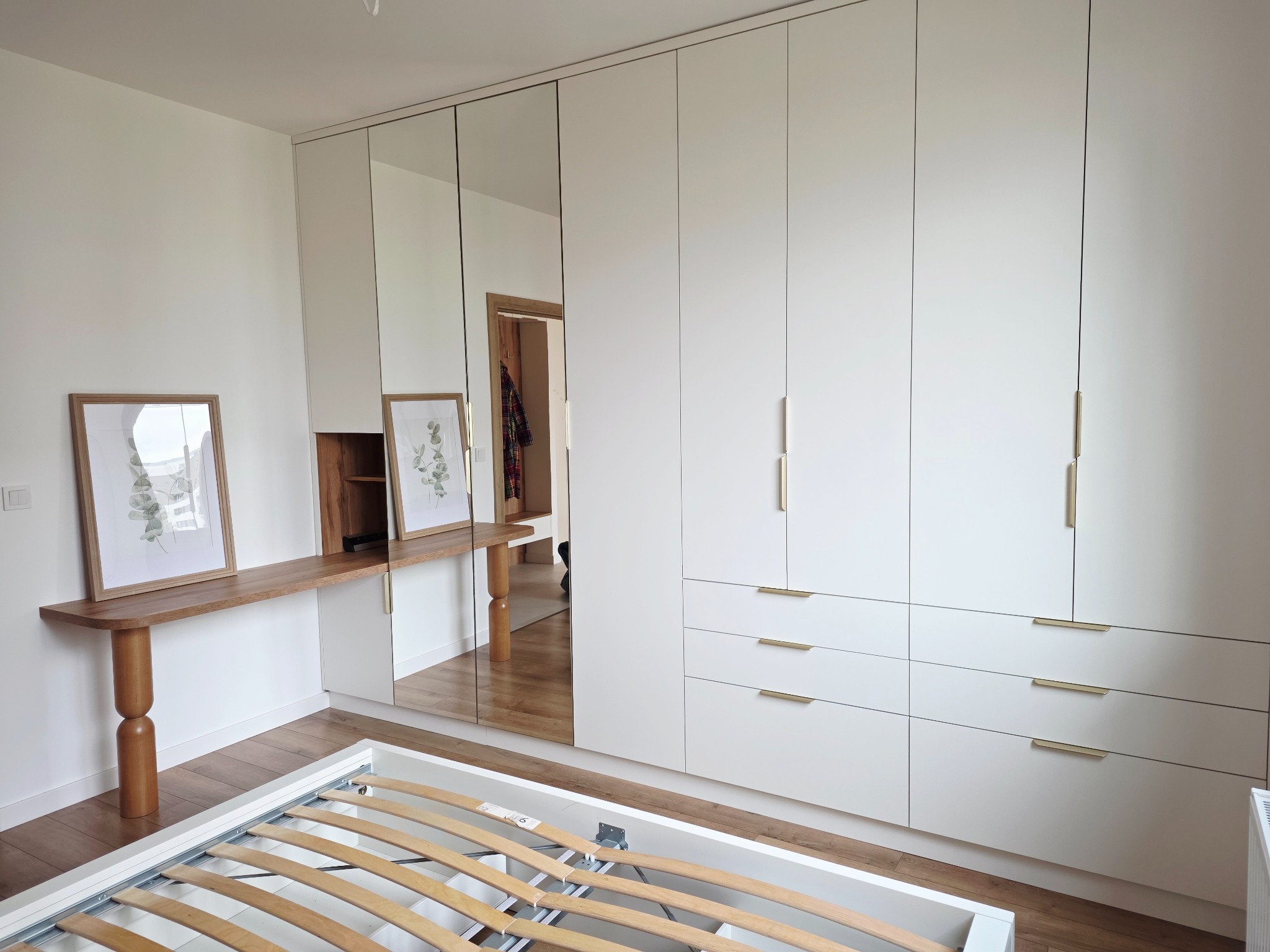 Biała szafa wnękowa z lustrzanymi drzwiami i drewnianym blatem z półką, uzupełniona o ramki ze zdjęciami. Widoczny stelaż łóżka. Minimalistyczny design.