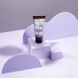 Fioletowa tuba peelingu do skóry głowy marki Be Natural, ustawiona na białych kostkach i fioletowych półokręgach na jasnofioletowym tle, w aranżacji studyjnej z wyraźnymi cieniami.