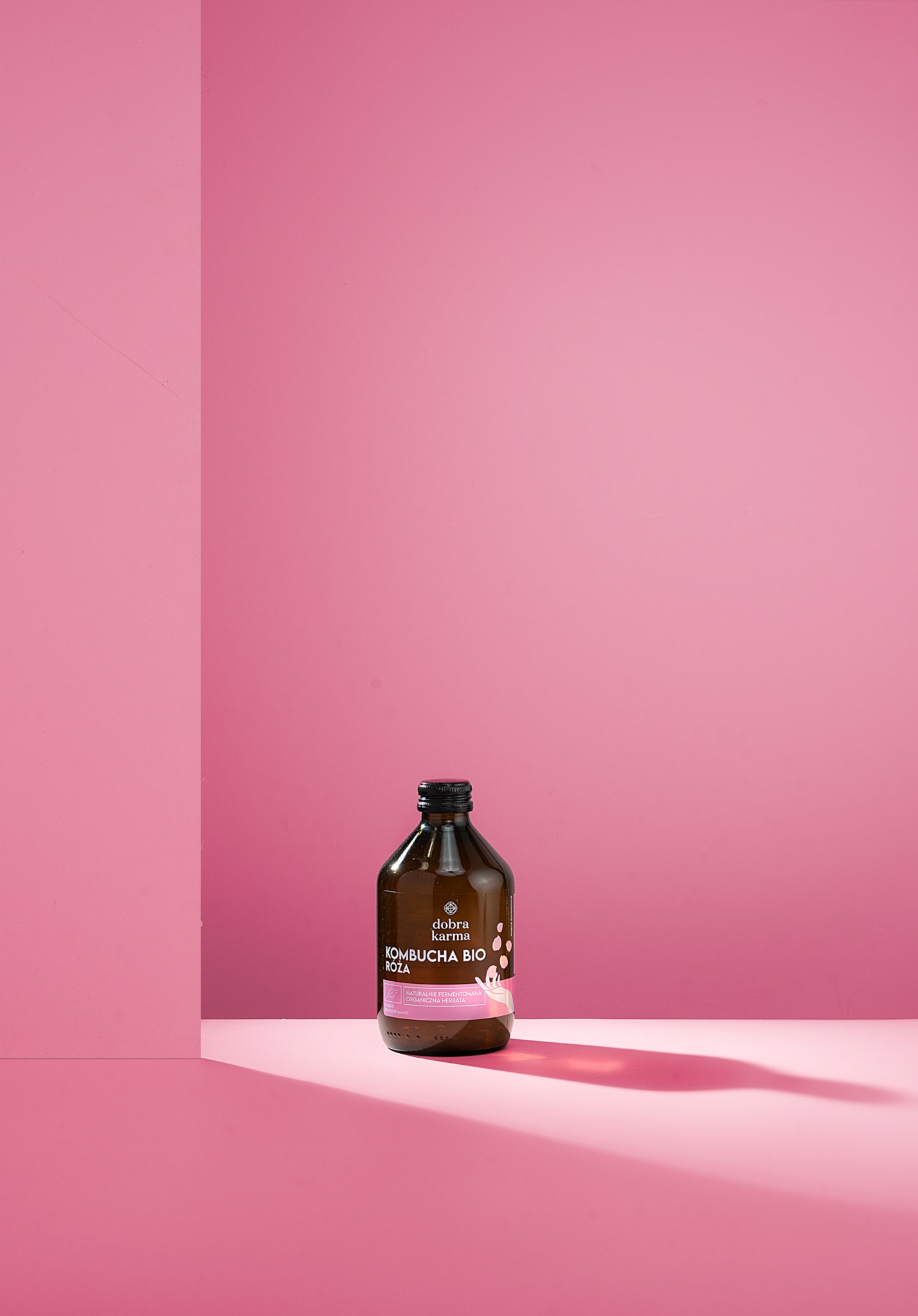 Butelka kombuchy bio róża marki Dobra Karma na różowym tle z wyraźnym cieniem, minimalistyczna kompozycja.
