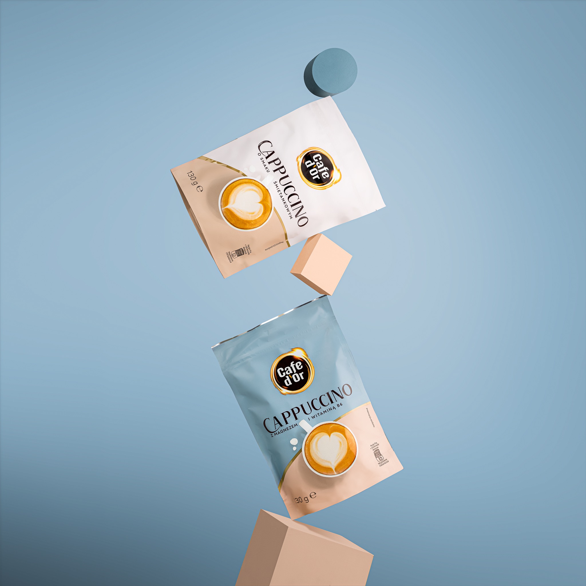 Dwa opakowania Cappuccino Cafe d'Or, jedno białe i jedno błękitne, unoszą się w powietrzu na tle błękitnego tła, otoczone geometrycznymi kształtami w kolorze beżowym i niebieskim.