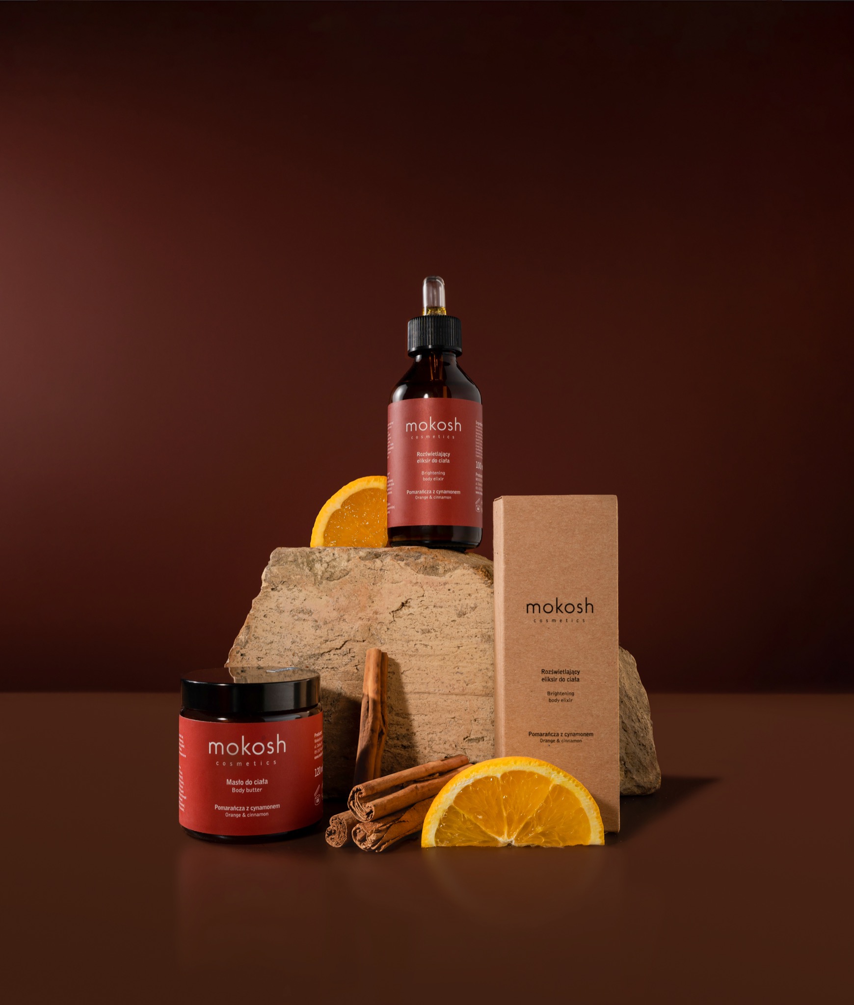 Kompozycja trzech kosmetyków Mokosh: masło do ciała, eliksir w butelce z pipetą i eliksir w kartonowym opakowaniu, uzupełniona o plaster pomarańczy i laski cynamonu, na tle w odcieniach brązu.