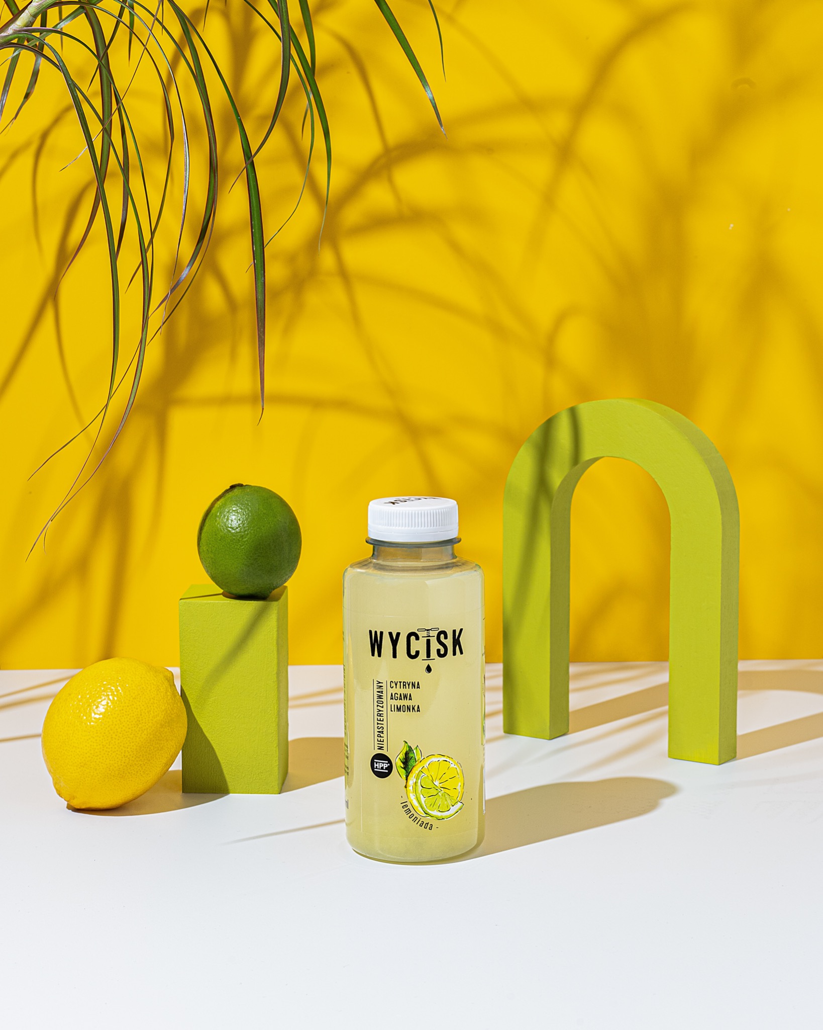 Stylizowana fotografia produktowa napoju Wycisk: lemoniada w plastikowej butelce, limonka na zielonym postumencie, cytryna obok, żółte tło z cieniem liści palmy i zielony łuk.