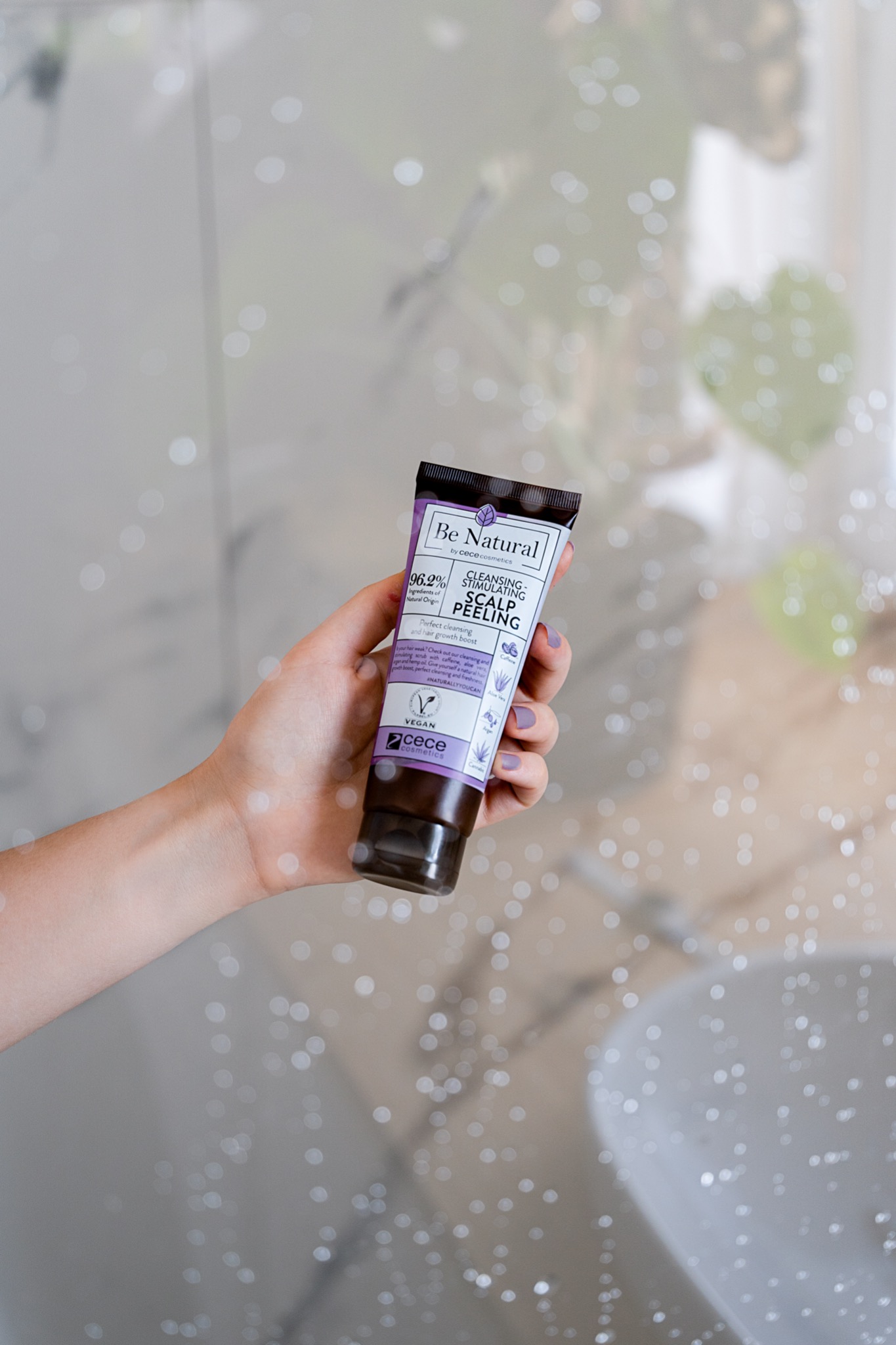 Dłoń z fioletowym manicure trzyma tubkę peelingu do skóry głowy marki Be Natural na tle rozmytej łazienki z umywalką i dekoracyjnymi światłami.