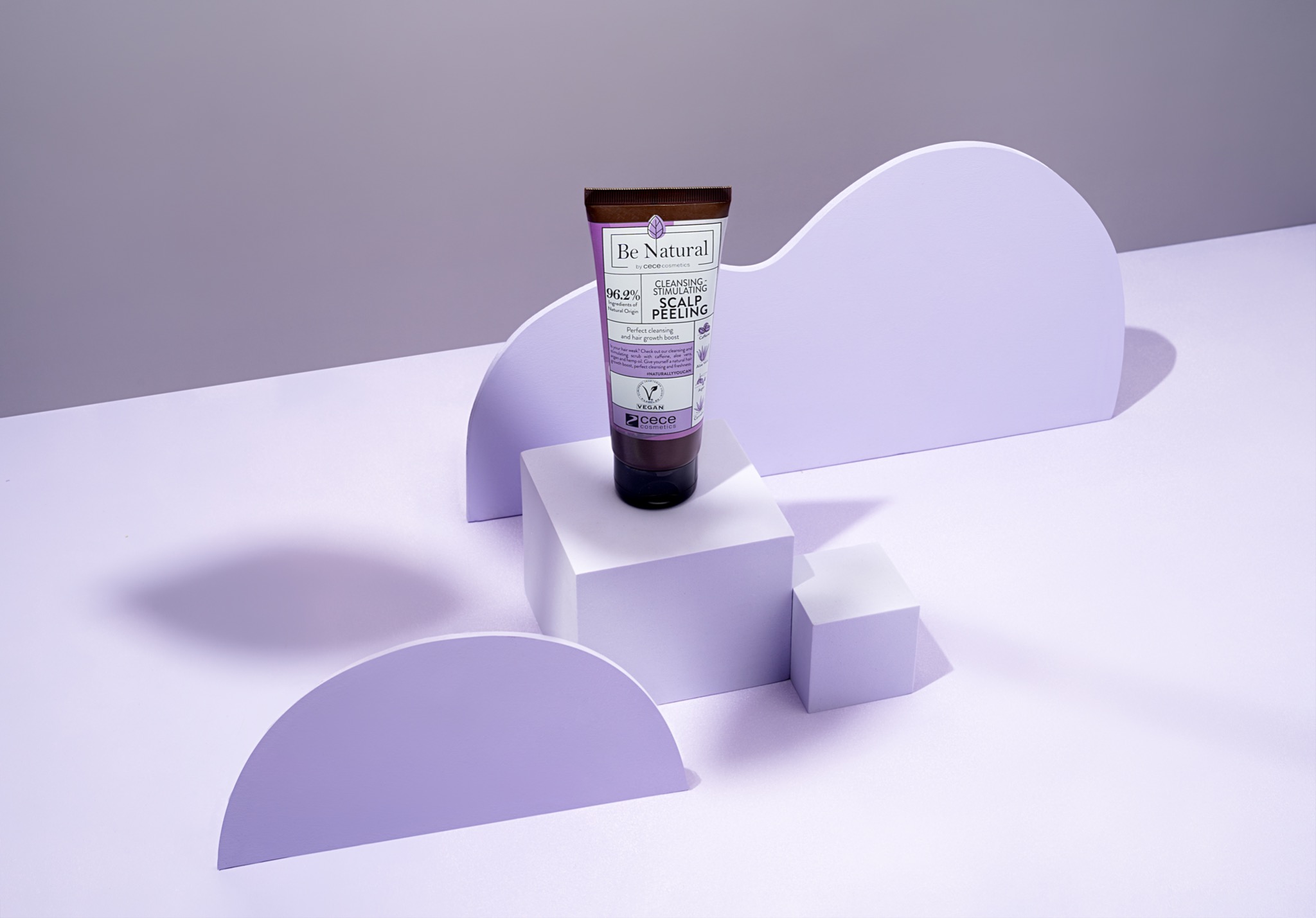 Fioletowa tuba peelingu do skóry głowy marki Be Natural, ustawiona na białych kostkach i fioletowych półokręgach na jasnofioletowym tle, w aranżacji studyjnej z wyraźnymi cieniami.