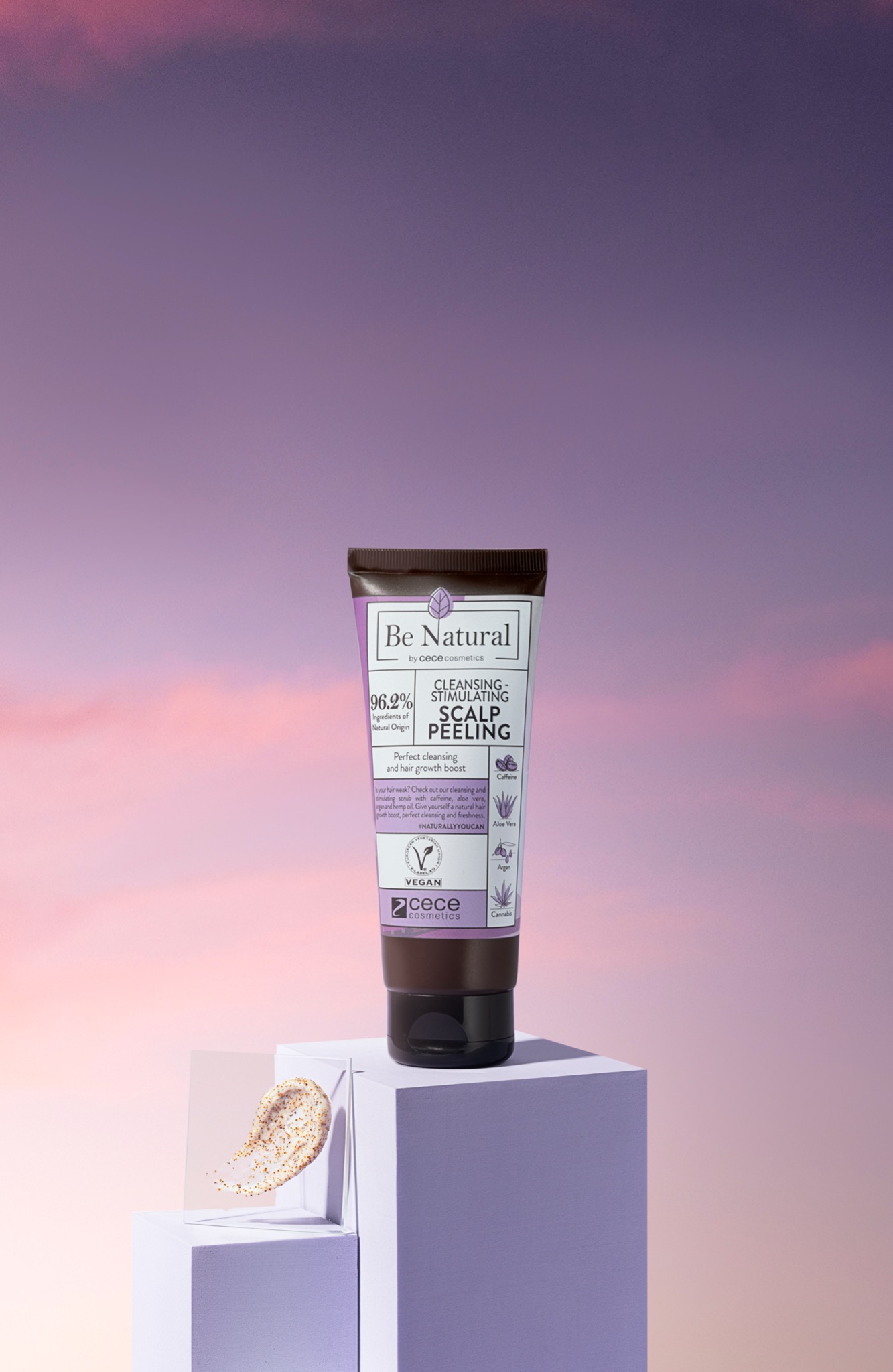 Tubka peelingu do skóry głowy marki Cece Cosmetics z linii Be Natural, umieszczona na fioletowym postumencie na tle pastelowego nieba o zmierzchu, obok próbka peelingu na przezroczystej płytce.