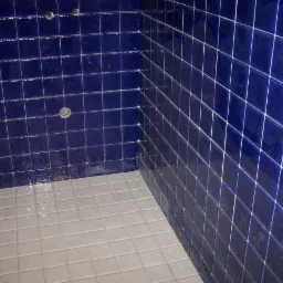 Niebieska glazura ścienna w kabinie prysznicowej z szarą podłogą. Widoczne otwory na armaturę. Ułożenie płytek w regularną siatkę z białą fugą.