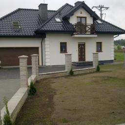 Webbud Wojciech Białachowski - Nowo wybudowany dom jednorodzinny z ciemną dachówką, białymi ścianami i drewnianymi elementami wykończeniowymi, widoczny z zewnątrz, z częściowo zagospodarowanym terenem.