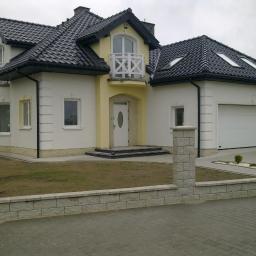 Webbud Wojciech Białachowski - Gotowy dom jednorodzinny z garażem, wykończony w kolorach białym i żółtym, z ciemną dachówką i elementami dekoracyjnymi na elewacji, otoczony niskim murkiem z szarej kostki.