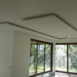 Nowoczesne wykończenie wnętrza: białe ściany, duże okna przesuwne z widokiem na zieleń, podwieszany sufit z dekoracyjnym oświetleniem. Minimalistyczny design.