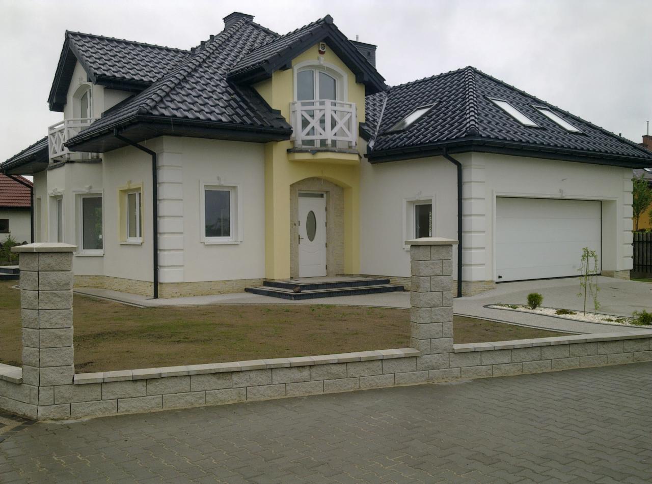 Gotowy dom jednorodzinny z garażem, wykończony w kolorach białym i żółtym, z ciemną dachówką i elementami dekoracyjnymi na elewacji, otoczony niskim murkiem z szarej kostki.