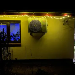 Zewnętrzna jednostka klimatyzacji Rotenso zamontowana na elewacji budynku, oświetlona ciepłym światłem lampek choinkowych, z widocznym oknem z niebieską poświatą w tle, sfotografowana nocą.