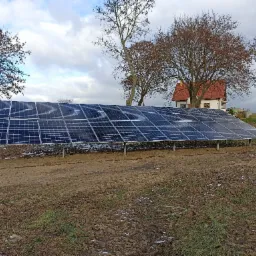 Instalacja paneli fotowoltaicznych na metalowej konstrukcji, widok na dom z czerwoną dach&oacute;wką i drzewa bez liści w tle, pochmurne niebo.