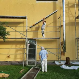 Pracownicy na rusztowaniach podczas prac elewacyjnych na tle żółtej ściany budynku z kominem, jeden z nich podnosi element rusztowania, a drugi obserwuje.