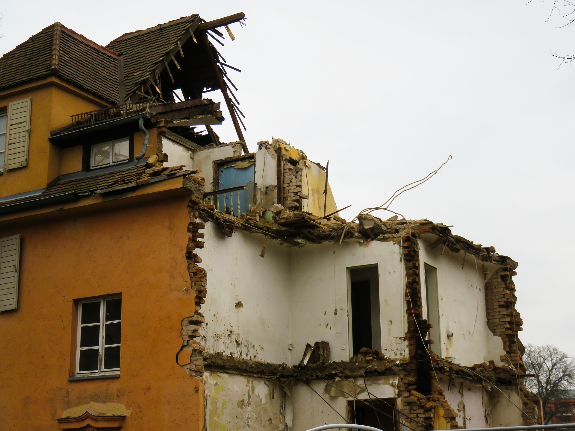 Częściowo zburzony, dwupiętrowy budynek z odsłoniętymi cegłami i fragmentami dachu, ukazujący zniszczone wnętrze z niebieskimi balustradami balkonu.