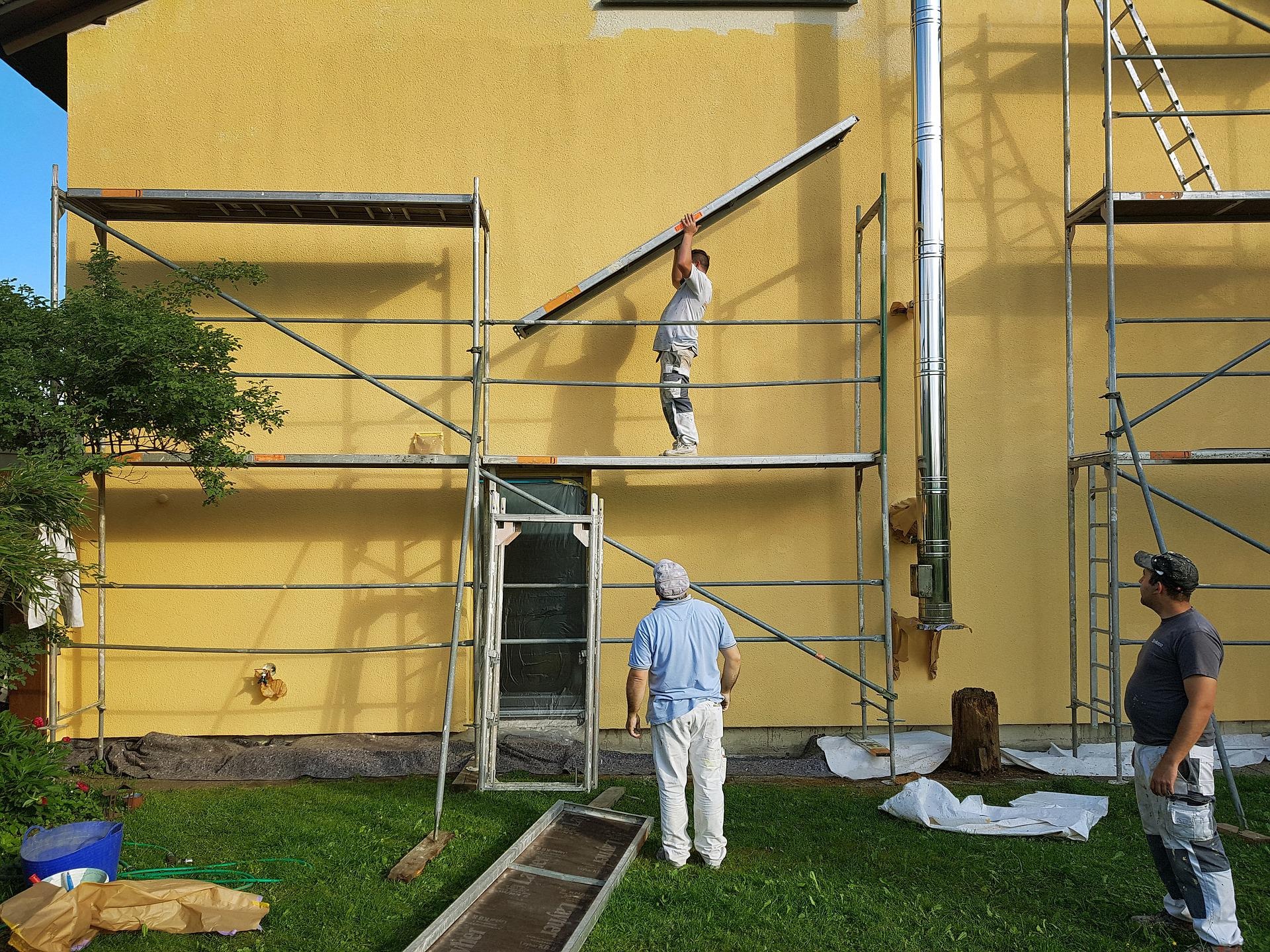Pracownicy na rusztowaniach podczas prac elewacyjnych na tle żółtej ściany budynku z kominem, jeden z nich podnosi element rusztowania, a drugi obserwuje.