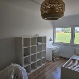 ARP-BUD Artur Pawlak - Wnętrze pokoju po odświeżeniu: białe ściany, drewniana podłoga, biały regał z kwadratowymi półkami, biurko z lampką, krzesło na kółkach, łóżko ze stelażem i widok na zielone pole za oknem.