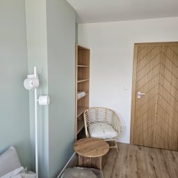 ARP-BUD Artur Pawlak - Jasne wnętrze pokoju z pomalowanymi na zielono ścianami, drewnianą podłogą i drzwiami z wzorem w jodełkę; widoczne meble: regał, wiklinowy fotel z poduszką, okrągły stolik, lampa oraz zapakowane...