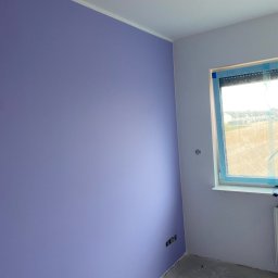 ARP-BUD Artur Pawlak - Pokój po świeżym malowaniu ścian na fioletowo, widoczne gniazdka elektryczne i okno z niebieską folią ochronną, surowa podłoga przed wykończeniem.