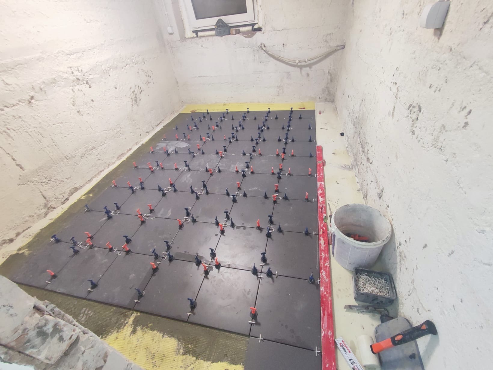 Układanie ciemnych płytek podłogowych z użyciem systemu poziomowania, widoczne krzyżyki dystansowe, wiadro i narzędzia przy ścianie z surowym tynkiem.