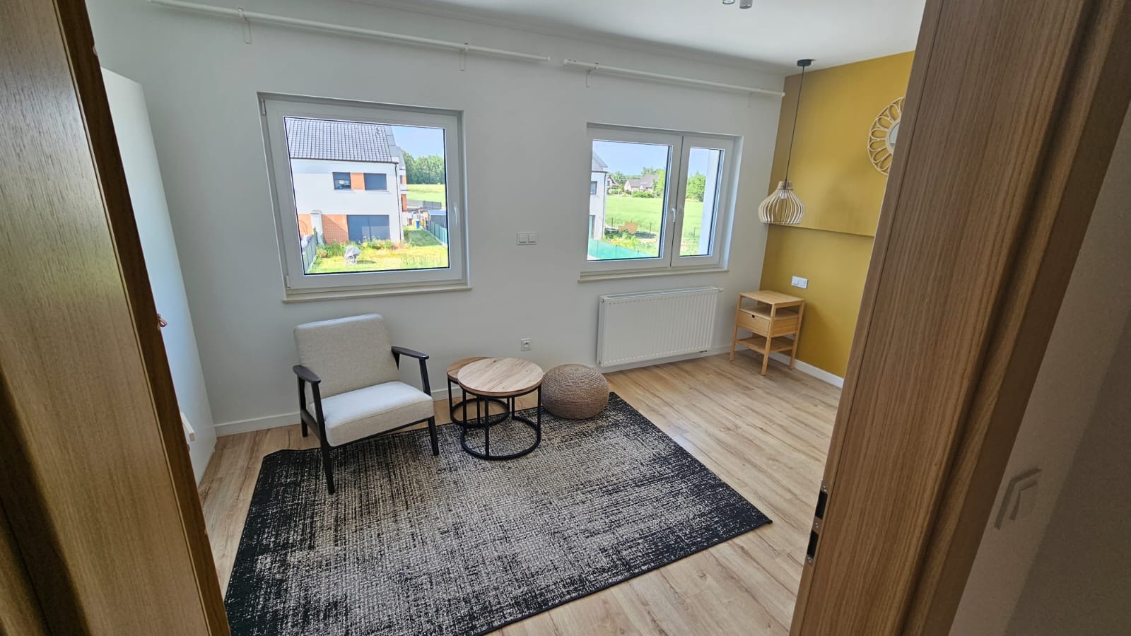 Jasny pokój z dwoma oknami, kremowym fotelem, okrągłymi stolikami, pufą i wzorzystym dywanem na drewnianej podłodze, ściana pomalowana na żółto w tle