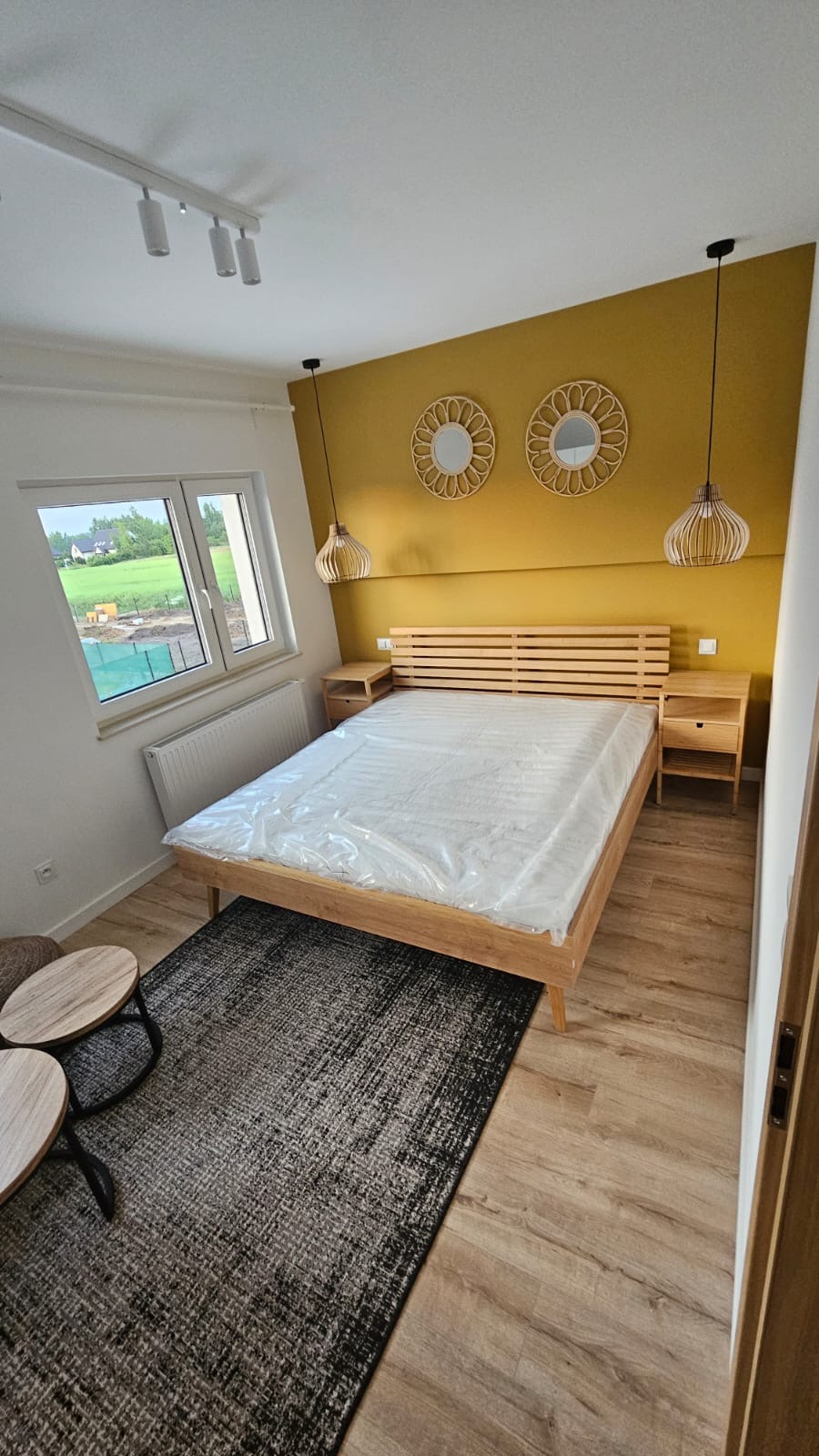 Jasne wnętrze sypialni z drewnianym łóżkiem z zagłówkiem w paski, nakastlikami, dywanem w odcieniach szarości, drewnianą podłogą, żółtą ścianą z okrągłymi lustrami i lampami wiszącymi, widokiem...