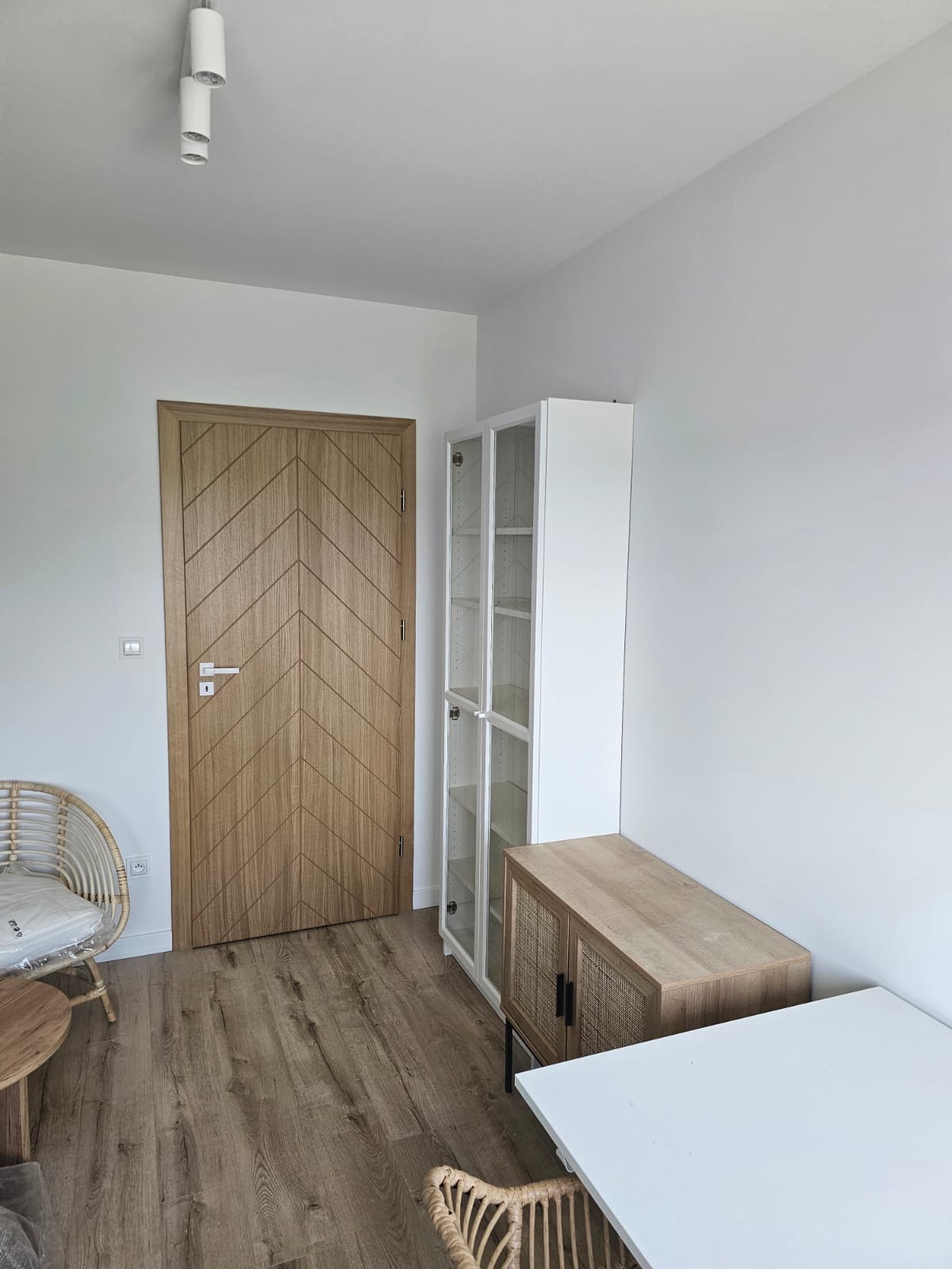 Jasne wnętrze mieszkania po remoncie: drewniana podłoga, białe ściany, drzwi z wzorem jodełki, biała witryna z przeszklonymi drzwiami, komoda z rattanowymi frontami i biały stół.