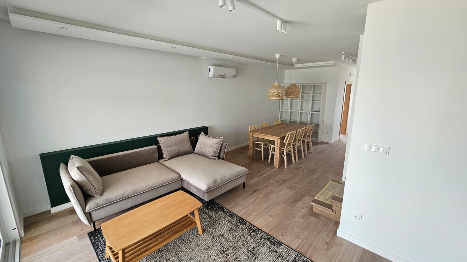 Jasne wnętrze salonu z drewnianą podłogą, beżową sofą narożną z poduszkami, drewnianym stolikiem kawowym na wzorzystym dywanie, stołem z krzesłami z rattanu pod plecionymi lampami i białą ścianą...