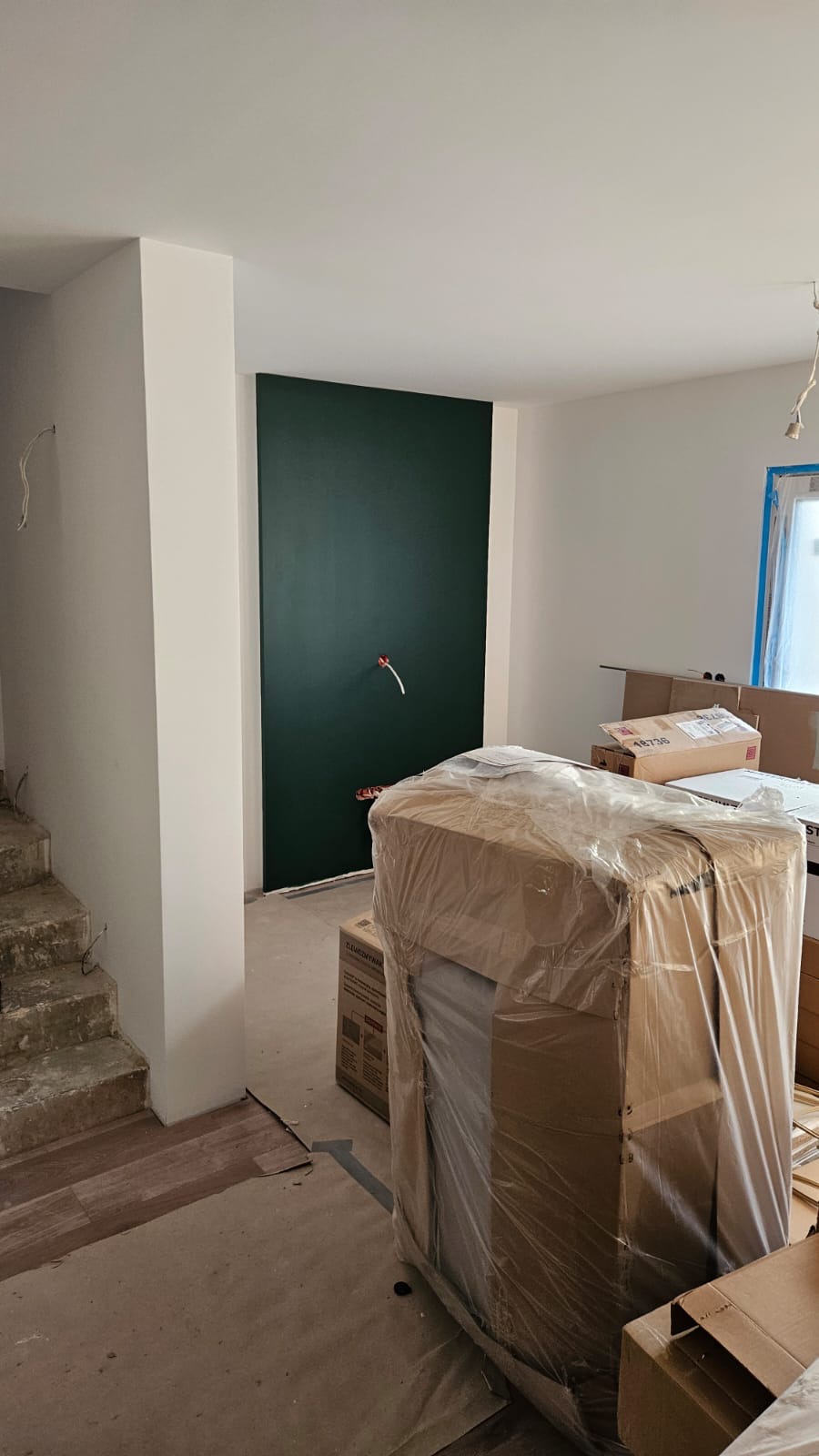 Wnętrze w trakcie wykańczania: ściana pomalowana na ciemnozielono z wystającymi instalacjami, częściowo ułożona podłoga z paneli, schody w stanie surowym oraz zapakowane elementy wyposażenia w folii.