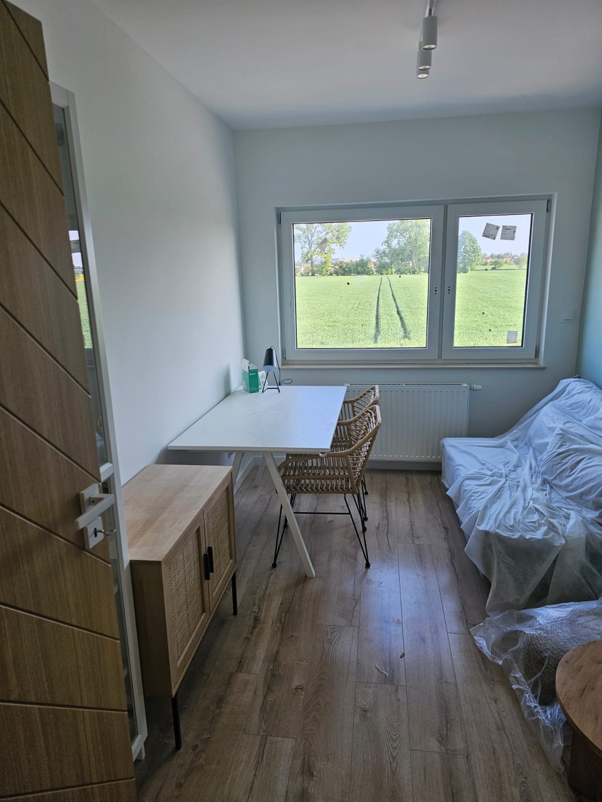 Jasne wnętrze pokoju po remoncie: świeżo pomalowane ściany, nowe panele podłogowe, biurko z krzesłami z rattanu, zabezpieczony folią mebel, widok na pole zza okna.