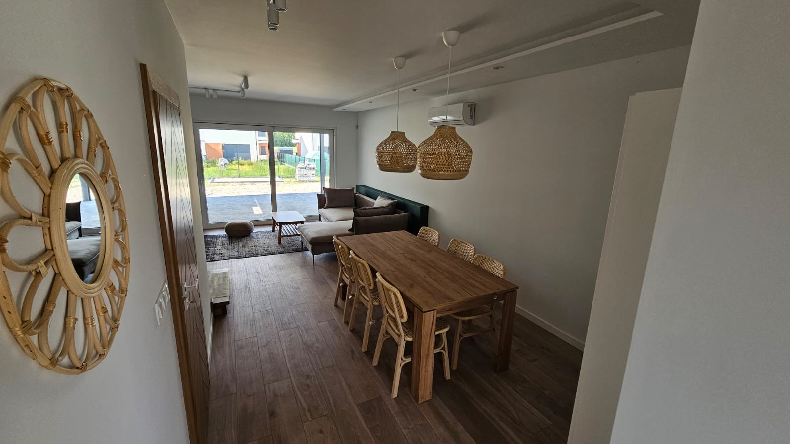 Jasne wnętrze salonu z drewnianą podłogą, dużym oknem przesuwnym, rattanowym lustrem na ścianie, drewnianym stołem i rattanowymi lampami wiszącymi.