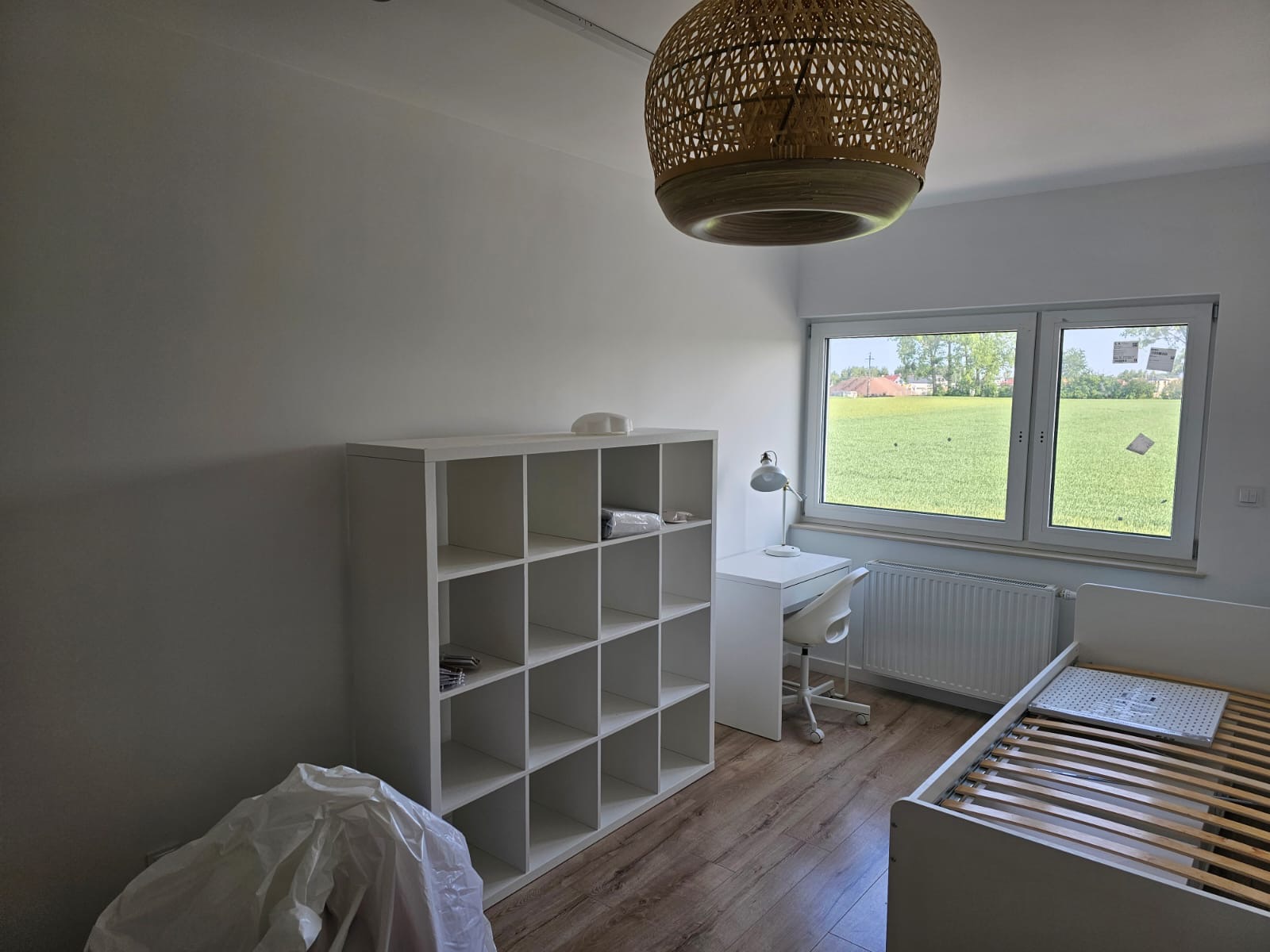 Wnętrze pokoju po odświeżeniu: białe ściany, drewniana podłoga, biały regał z kwadratowymi półkami, biurko z lampką, krzesło na kółkach, łóżko ze stelażem i widok na zielone pole za oknem.