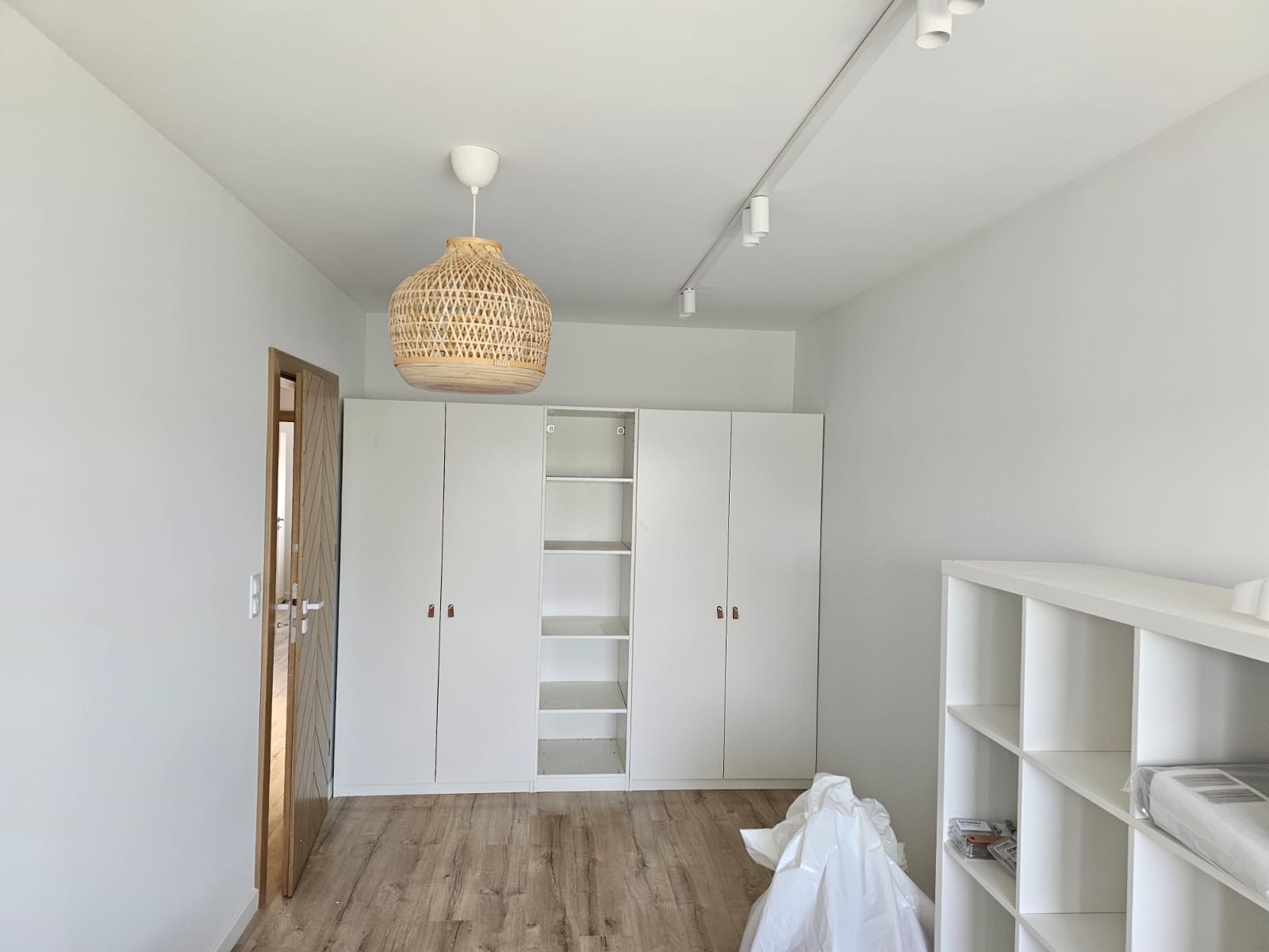 Jasne wnętrze pokoju po odświeżeniu, widoczne białe ściany, drewniana podłoga, białe szafy z brązowymi uchwytami, regał, lampa z wikliny oraz folia ochronna na podłodze, sugerująca niedawne prace...