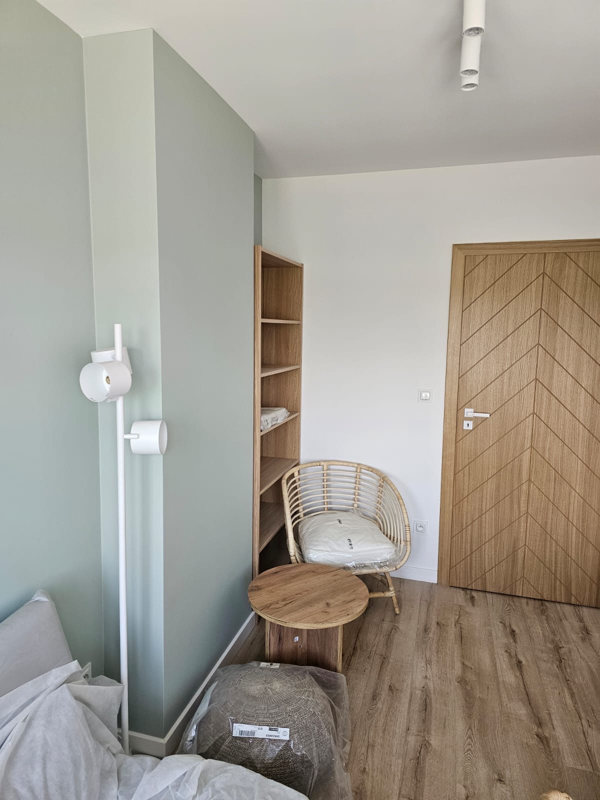 Jasne wnętrze pokoju z pomalowanymi na zielono ścianami, drewnianą podłogą i drzwiami z wzorem w jodełkę; widoczne meble: regał, wiklinowy fotel z poduszką, okrągły stolik, lampa oraz zapakowane...