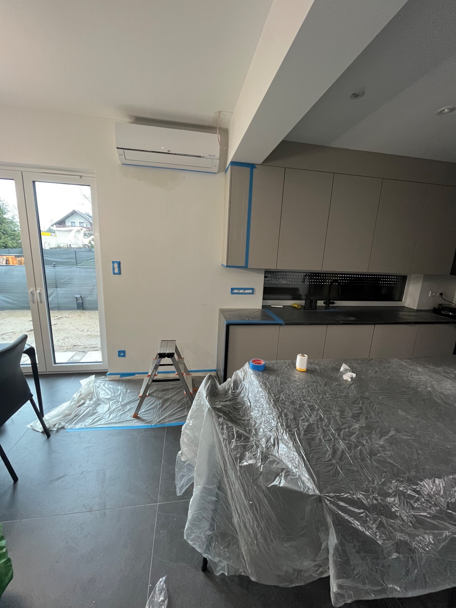 Przygotowania do prac malarskich w nowoczesnej kuchni: zabezpieczone folią meble i podłoga, drabina oparta o ścianę, taśma malarska na krawędziach, widoczny klimatyzator.
