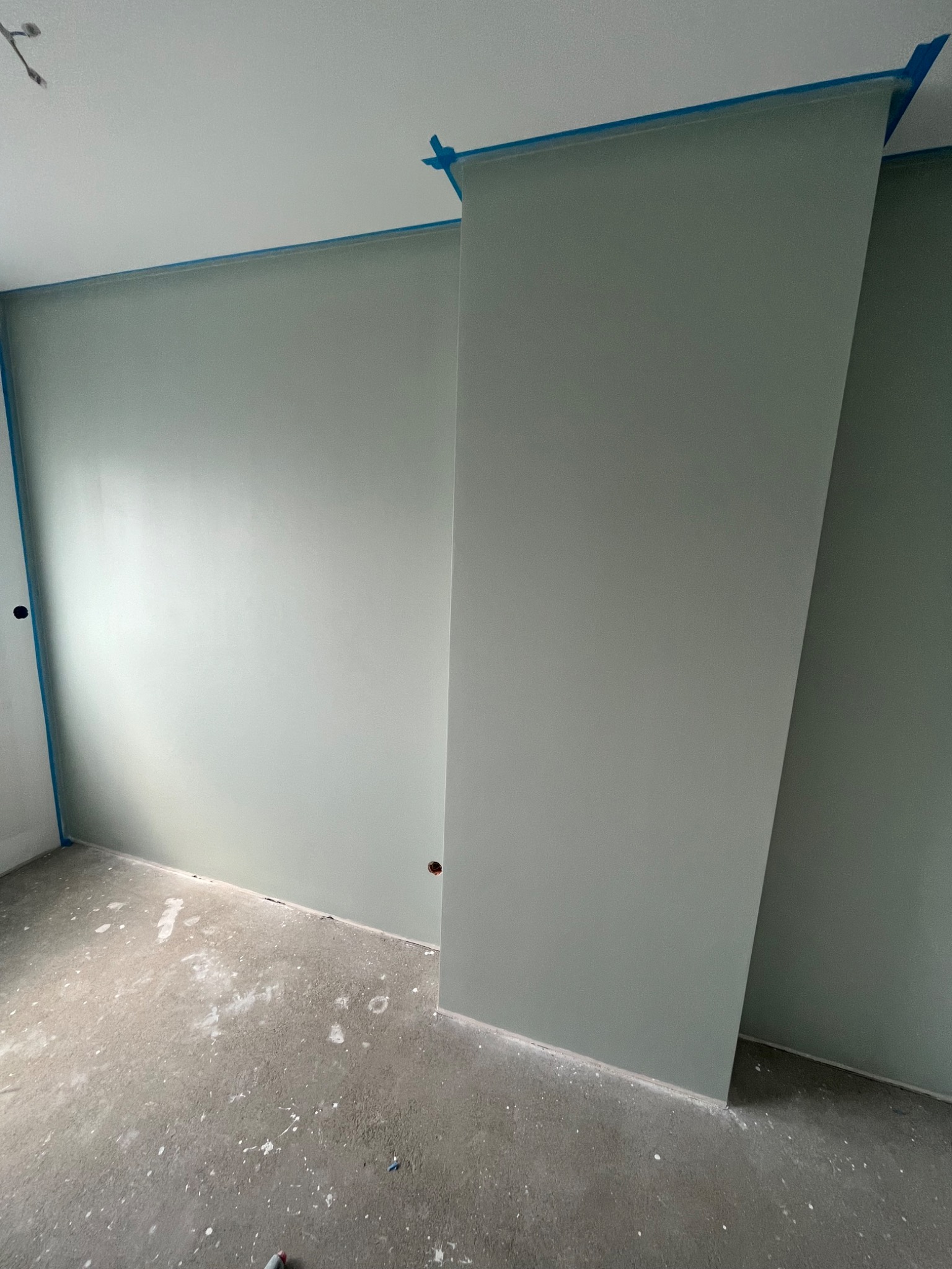 Świeżo pomalowane na szaro-zielono ściany w pomieszczeniu z zabezpieczonymi taśmą malarską krawędziami sufitu i ścian; podłoga surowa, z widocznymi plamami farby.
