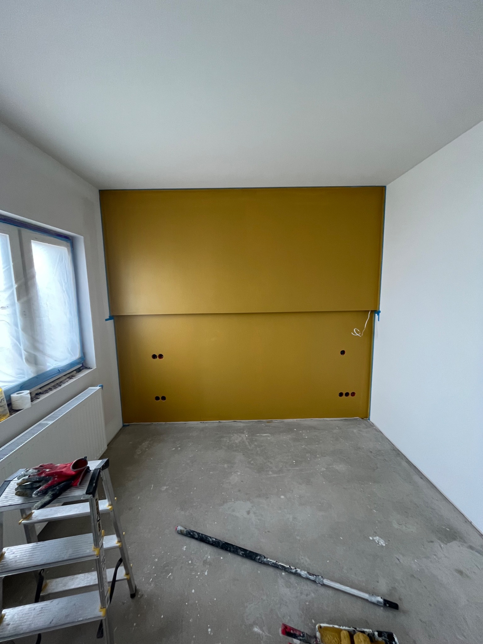 Pokój w trakcie malowania: jedna ściana pomalowana na musztardowy kolor, zabezpieczone taśmą listwy, widoczne puszki elektryczne, na podłodze drabina z rękawicami i wałek malarski.