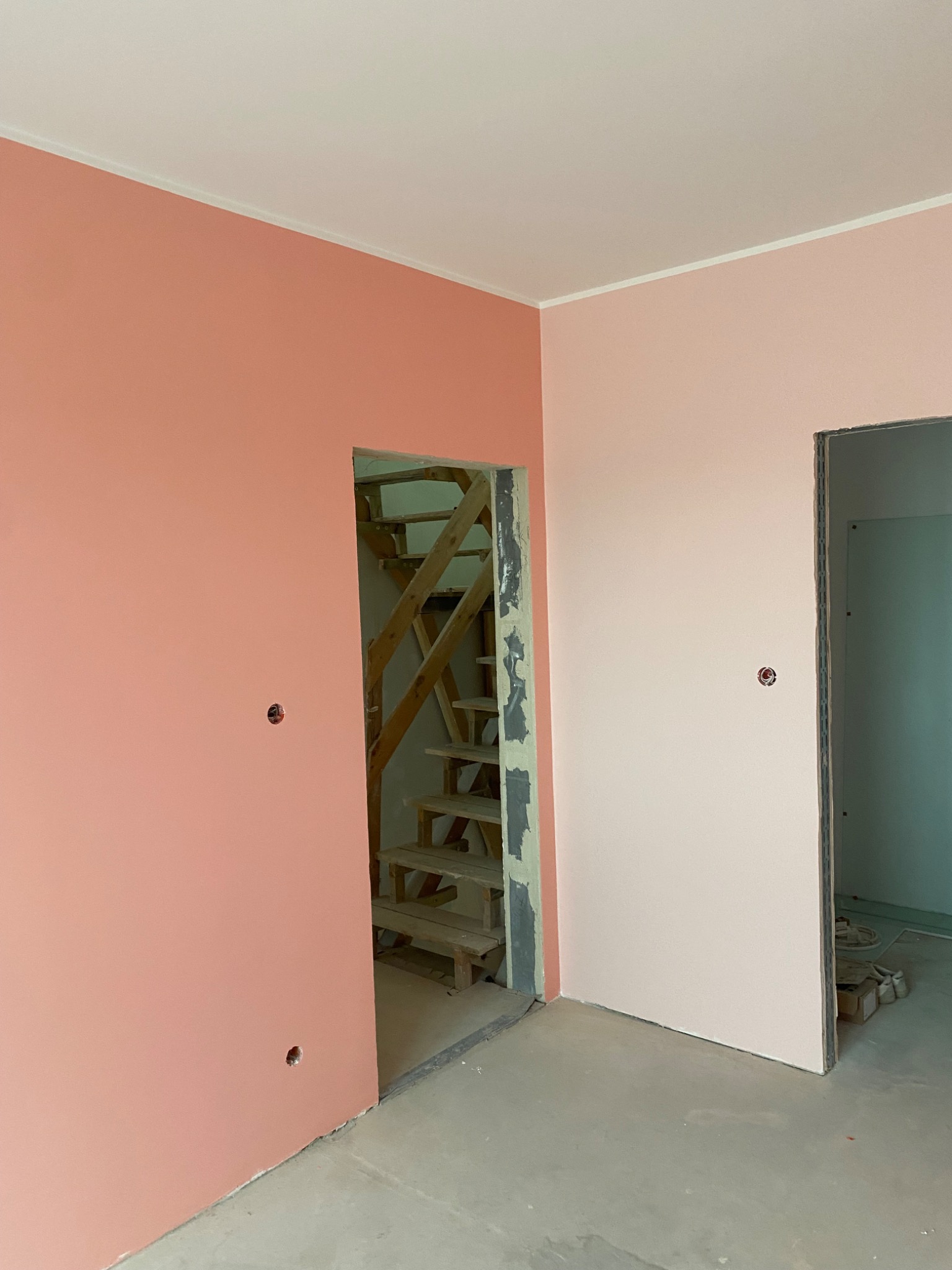Wnętrze w trakcie remontu: ściany pomalowane na dwa odcienie różu, widoczne otwory na gniazdka, drewniane schody w surowym stanie w prześwicie ściany, surowy beton na podłodze.