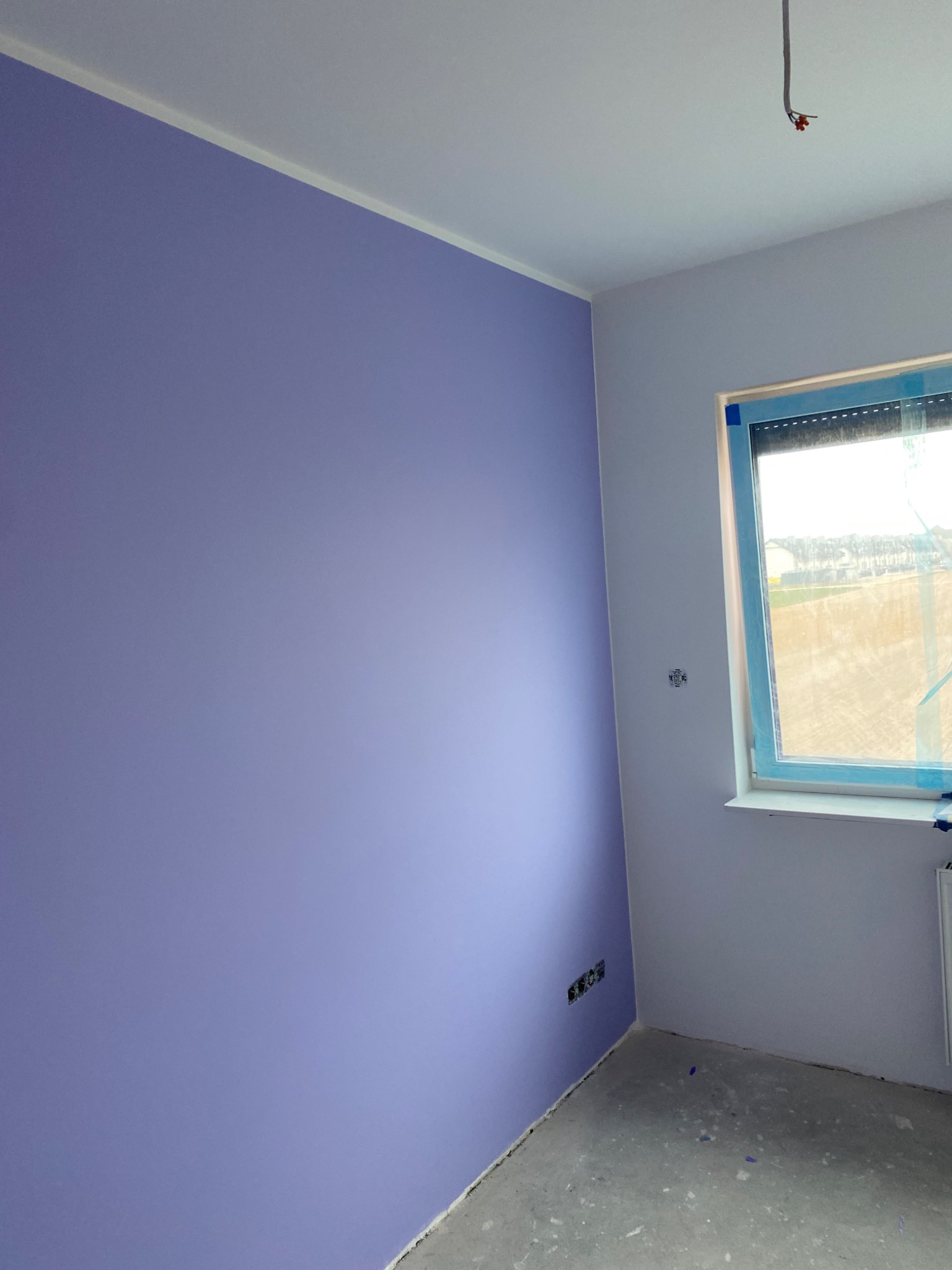 Pokój po świeżym malowaniu ścian na fioletowo, widoczne gniazdka elektryczne i okno z niebieską folią ochronną, surowa podłoga przed wykończeniem.