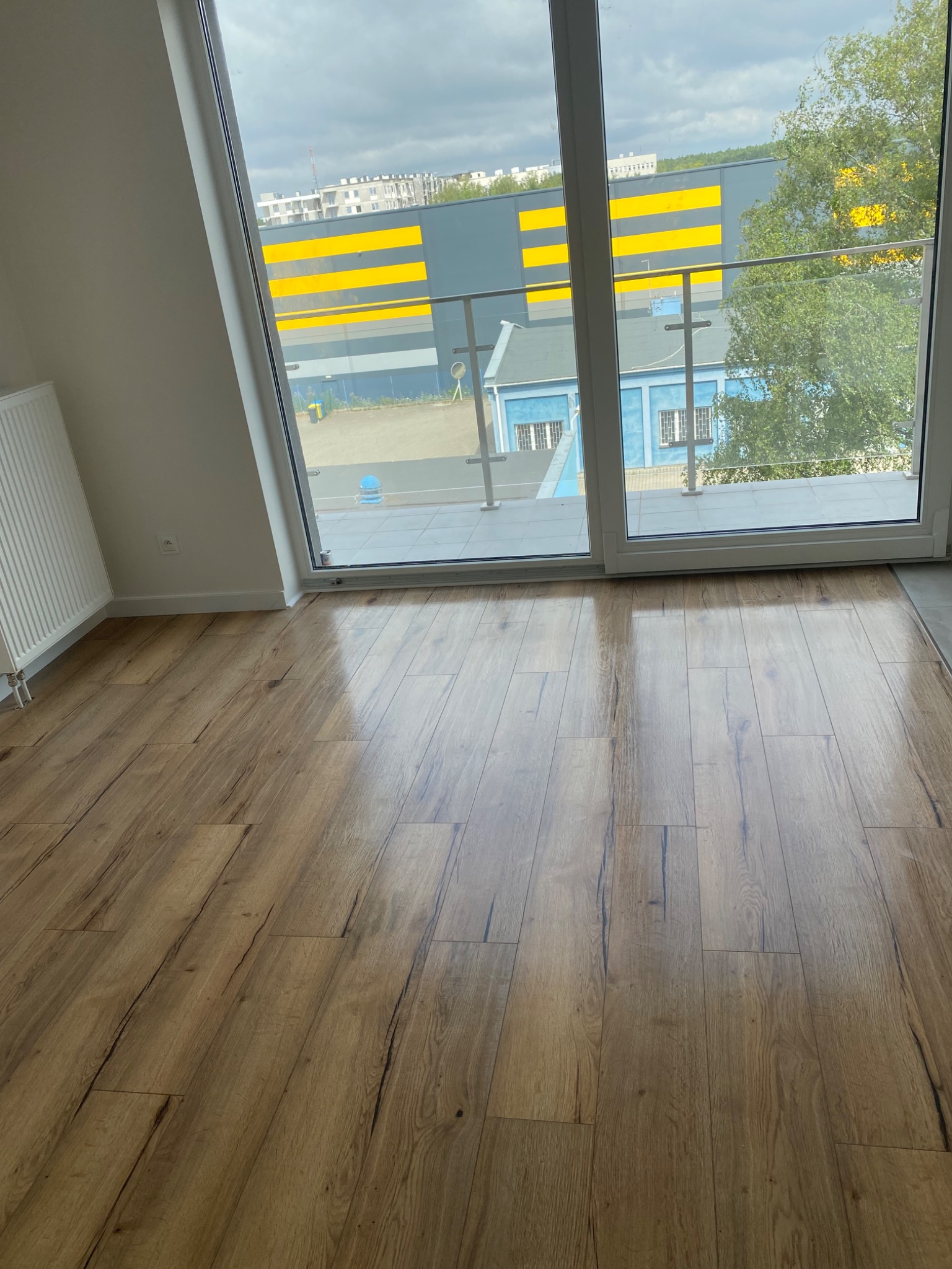 Świeżo ułożone panele podłogowe o wzorze drewna, widok z okna na budynek z żółtymi pasami, minimalistyczne wnętrze z białym grzejnikiem.