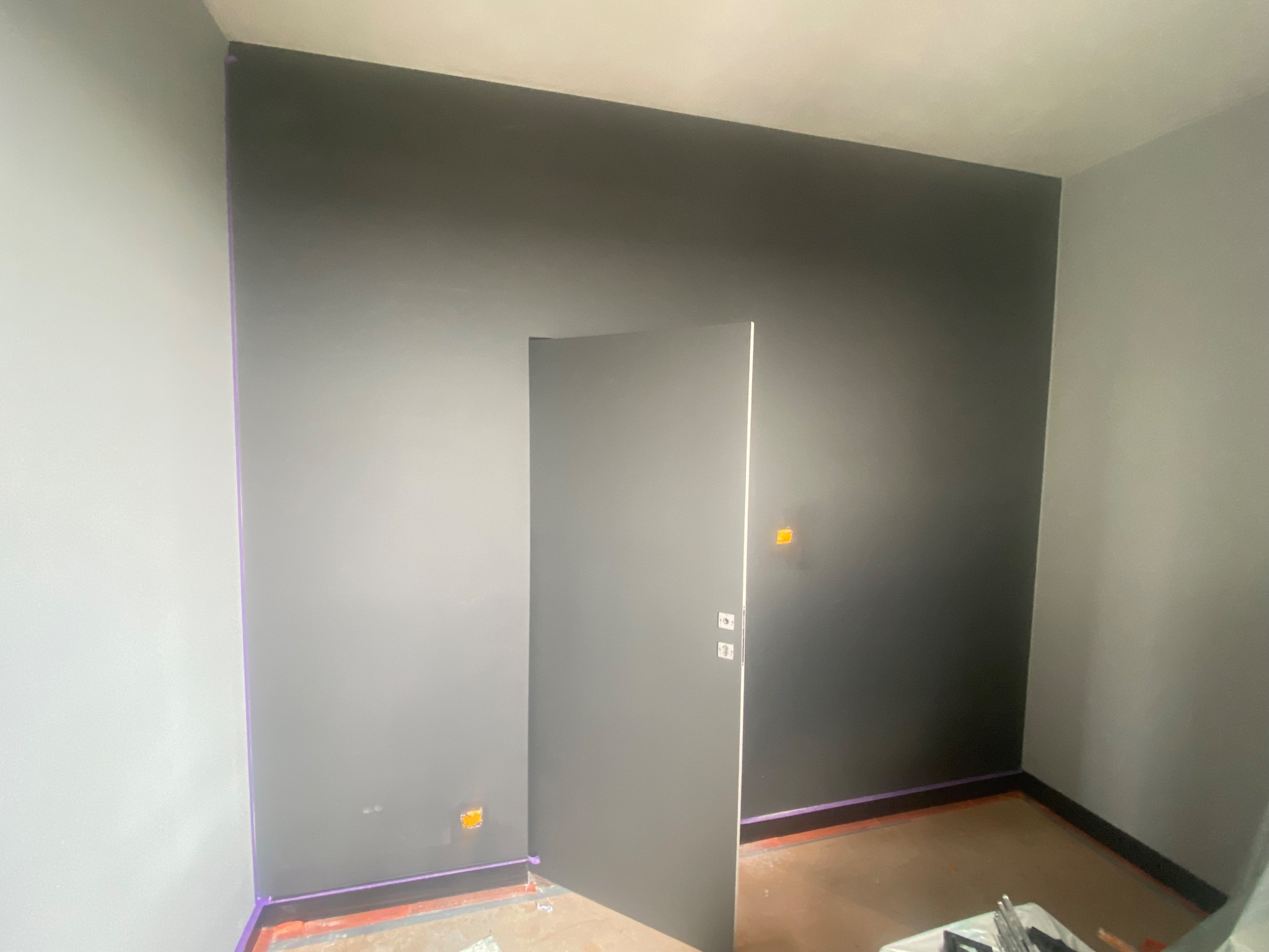 Pokój po malowaniu, ściany w odcieniach szarości, zabezpieczone taśmą malarską krawędzie, widoczne puszki elektryczne, częściowo otwarte drzwi.