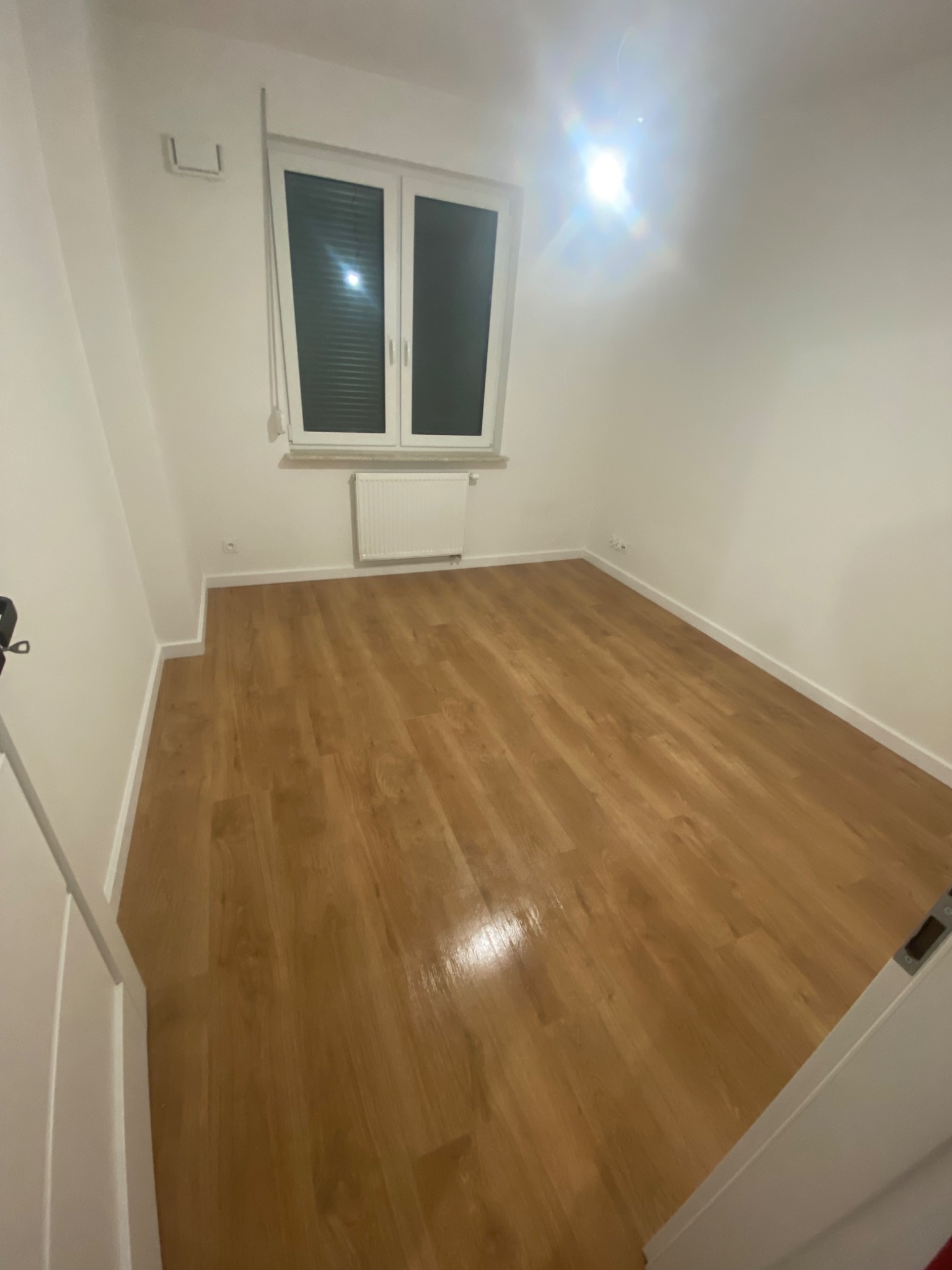 Pusty pokój z panelami podłogowymi w odcieniach brązu, białymi ścianami, oknem z żaluzjami i grzejnikiem. Widoczne listwy przypodłogowe. Światło słoneczne odbija się od powierzchni podłogi.