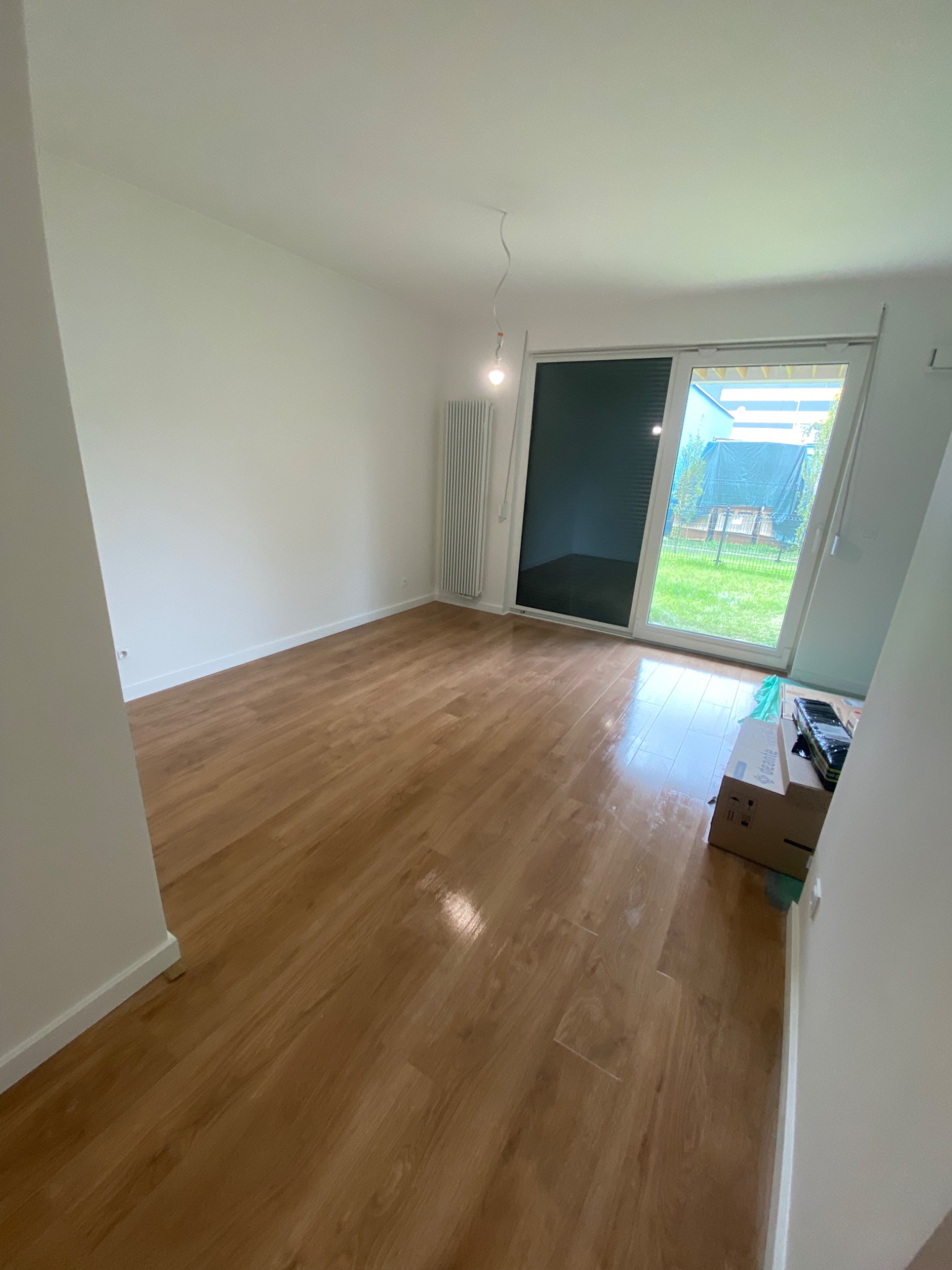 Pusty pokój w Poznaniu z parkietem, białymi ścianami i grzejnikiem, przygotowany do prac wykończeniowych, z widokiem na ogród przez przesuwne okno balkonowe i kartonami w rogu.