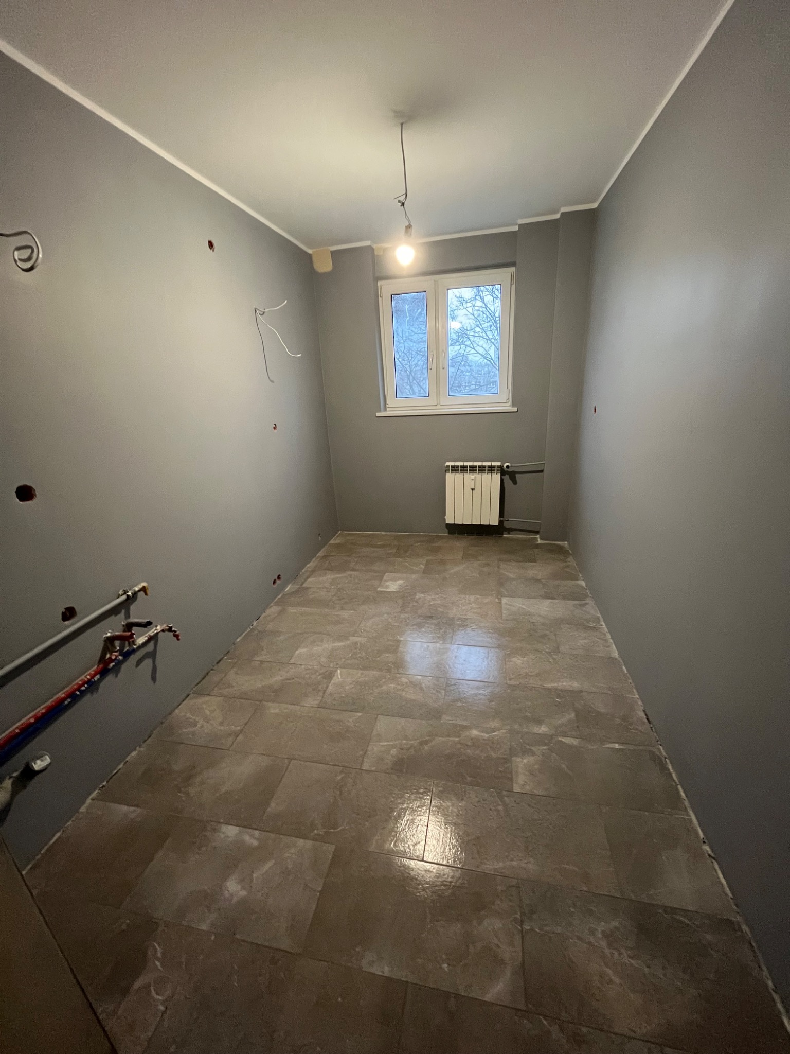Pokój po remoncie z szarymi ścianami, nową podłogą z płytek imitujących marmur i grzejnikiem pod oknem. Widoczne instalacje elektryczne i hydrauliczne w ścianach.