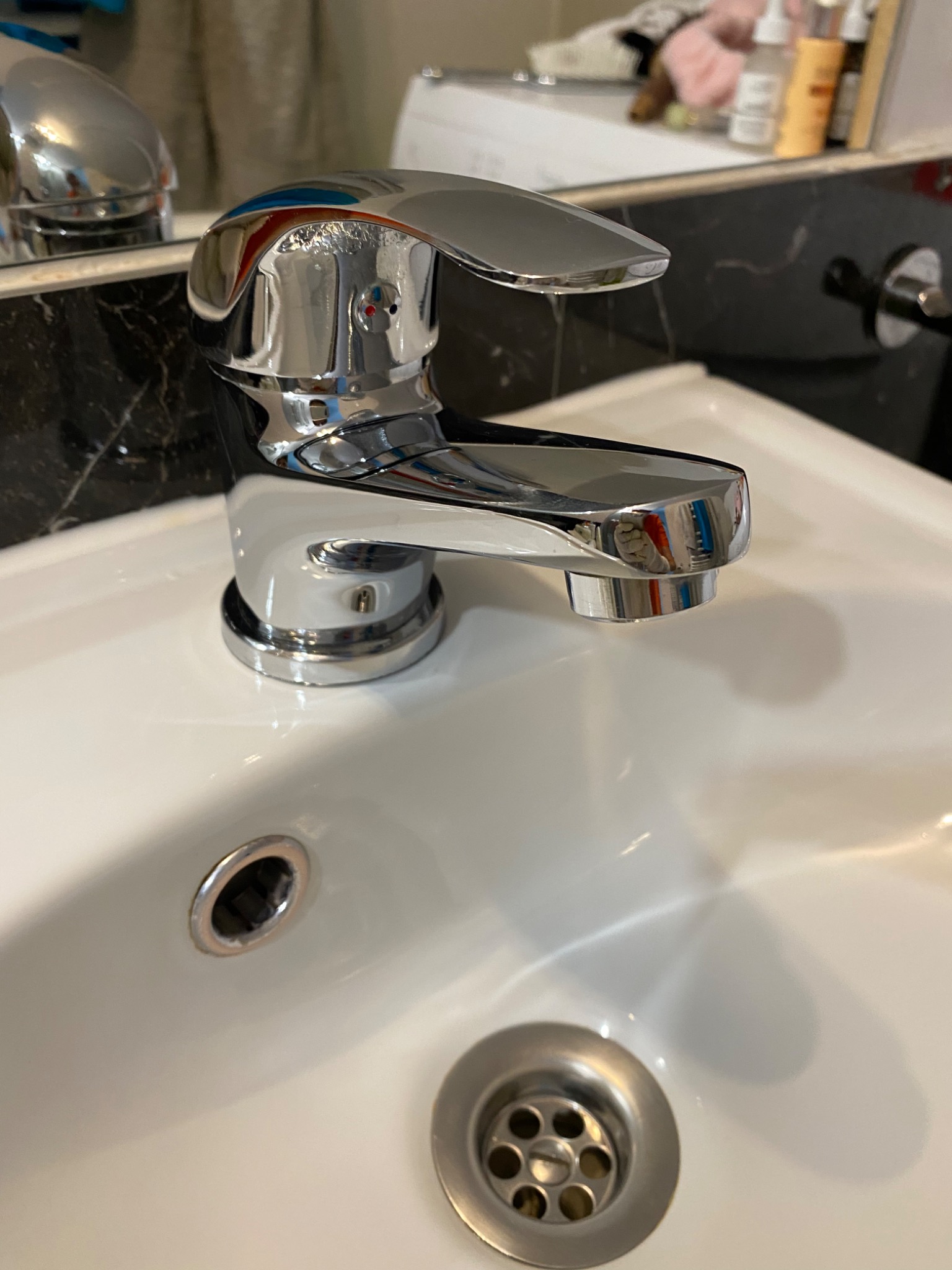 Błyszcząca, chromowana bateria umywalkowa z widocznymi śladami po kroplach wody na białej umywalce z metalowym odpływem.