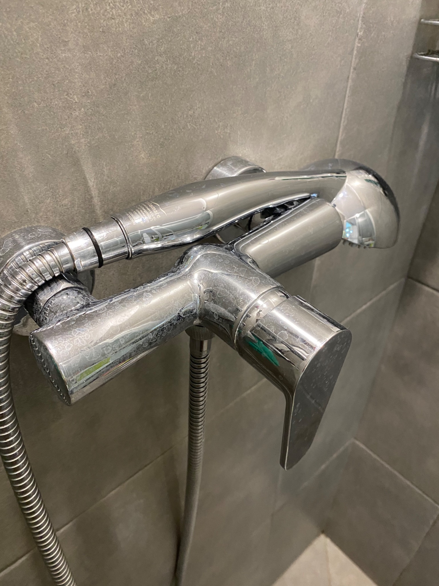 Bateria prysznicowa ze śladami kamienia na chromowanej powierzchni, zamontowana na szarej, kafelkowanej ścianie.